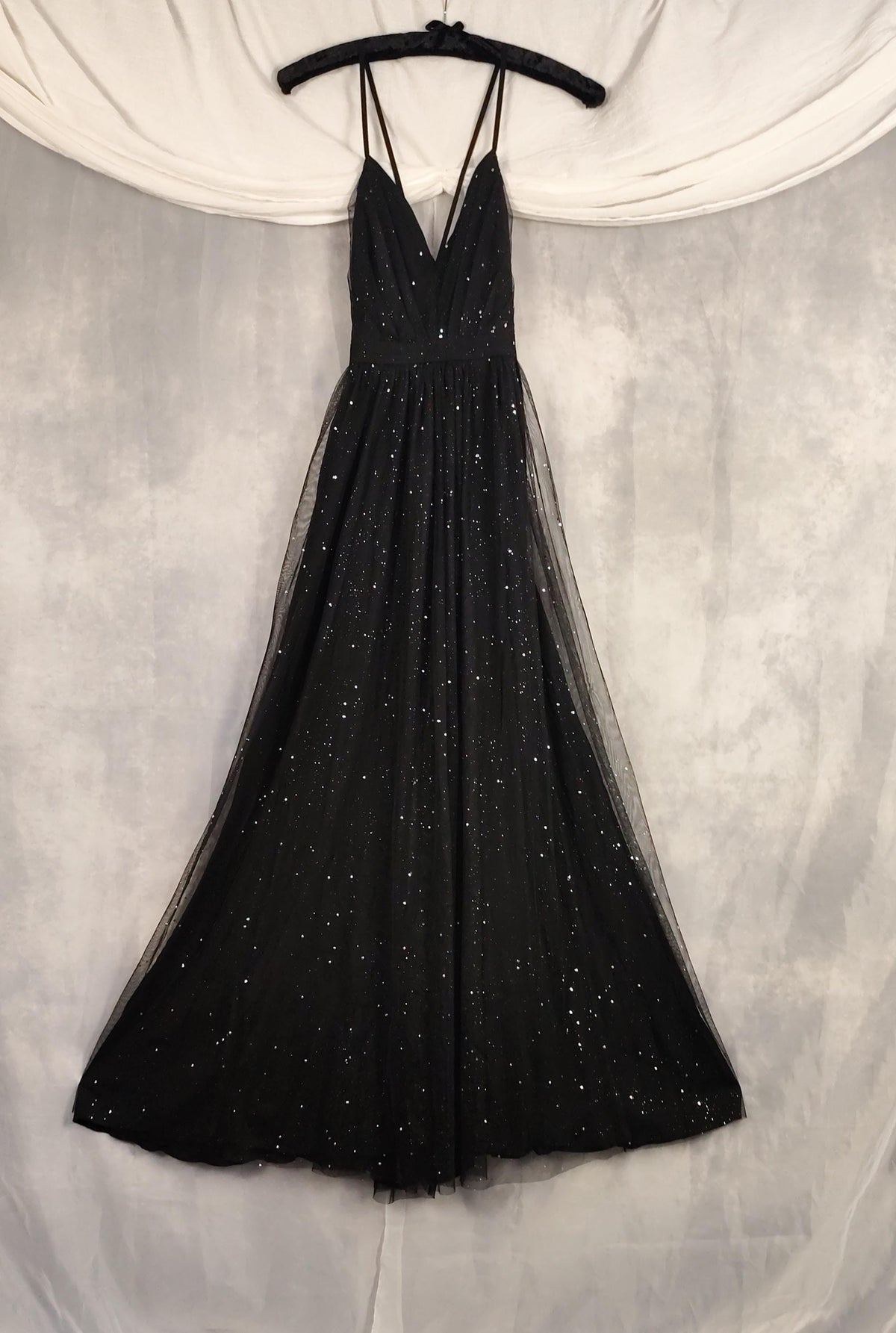 A black A-line evening gown with thin spaghetti straps and a V-neckline, decorated with sparkling sequins and tulle, hanging on a black hanger against a draped white fabric backdrop.
