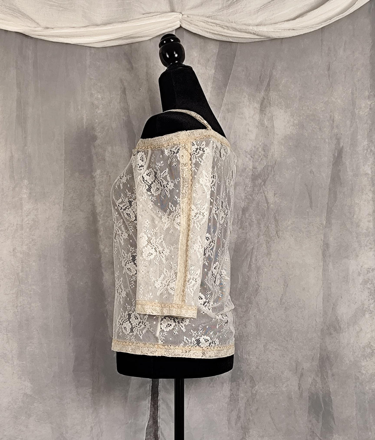 Side view of an off-the-shoulder lace top in cream and pearl, displayed on a black dress form mannequin against a textured gray backdrop with draped white fabric at the top.