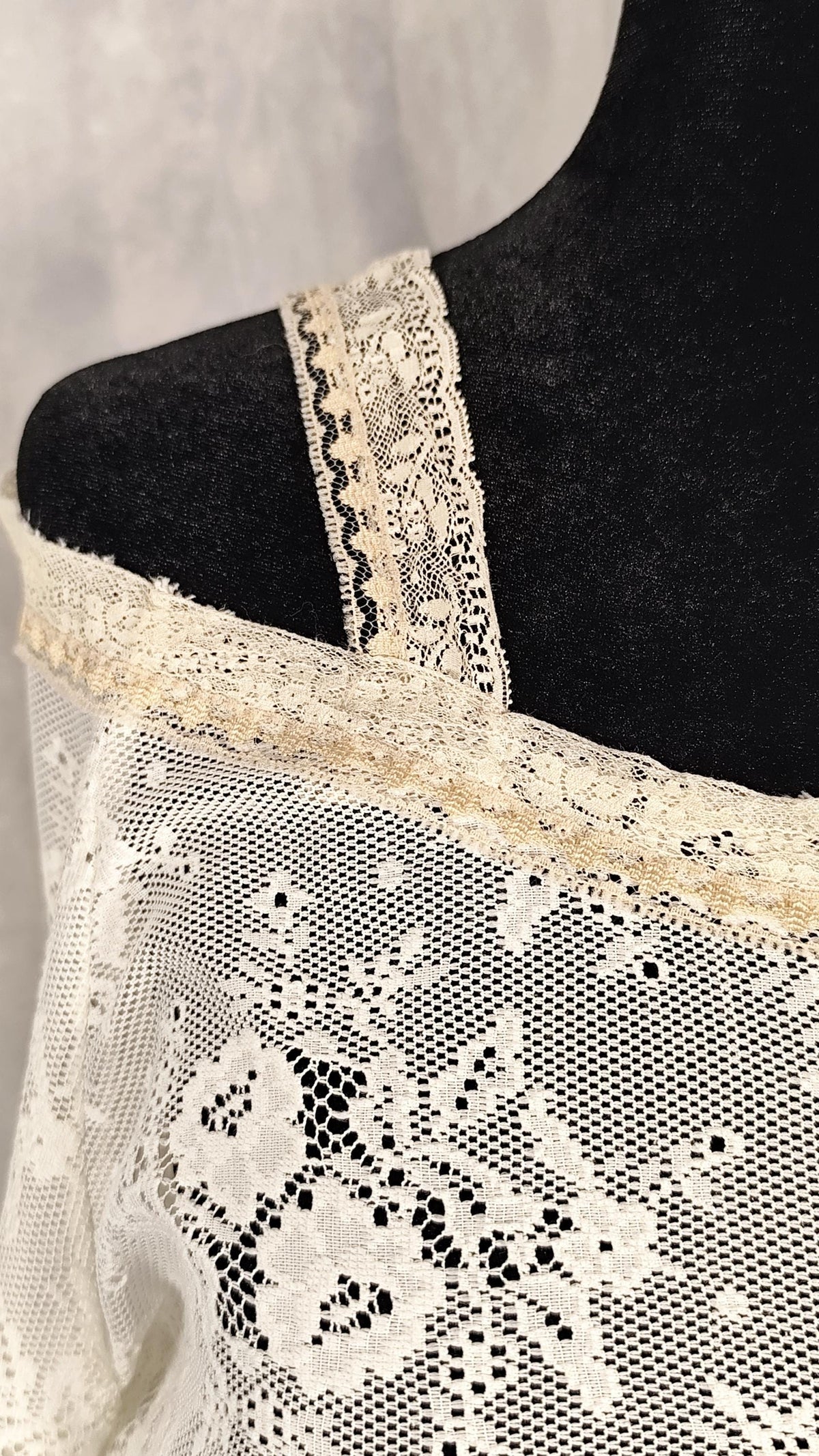 Close-up of a cream lace blouse draped on a black mannequin torso, showing floral lace fabric and wide lace trim against a light gray background.