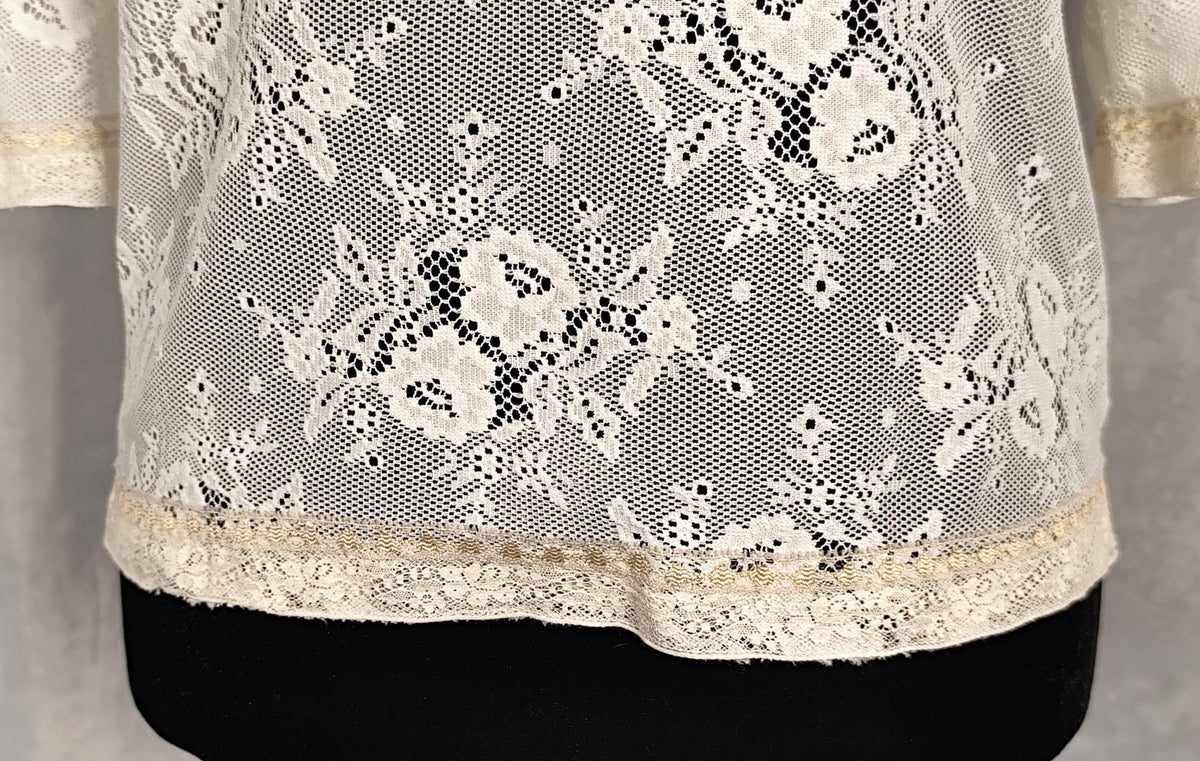 A pearl lace blouse with a floral pattern and a decorative lace hem, displayed on a black mannequin torso against a neutral gray background.