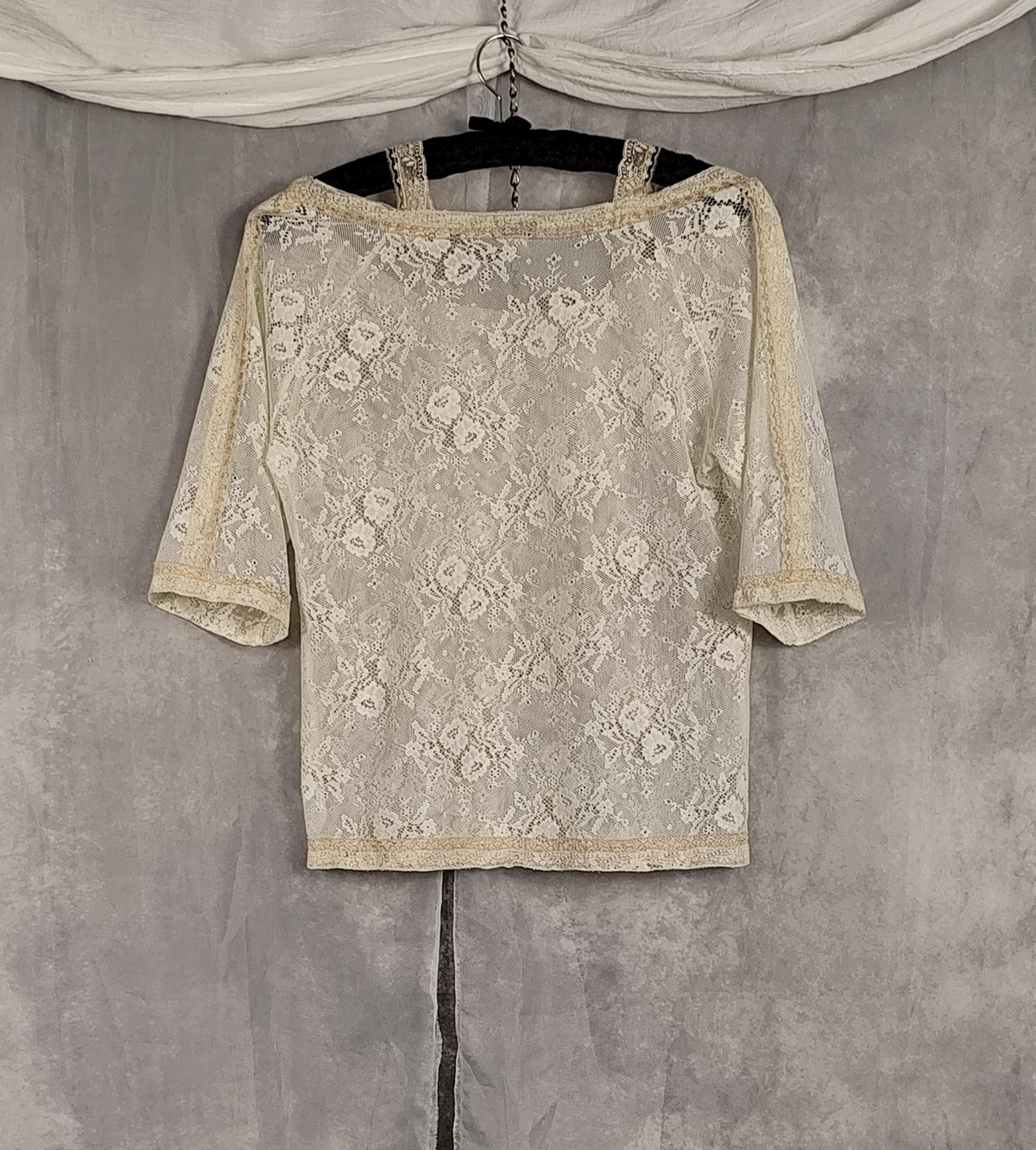 Back view of a pearl-colored lace 3/4 sleeve top with a floral pattern and lace trim, hanging on a black hanger against a textured gray backdrop.