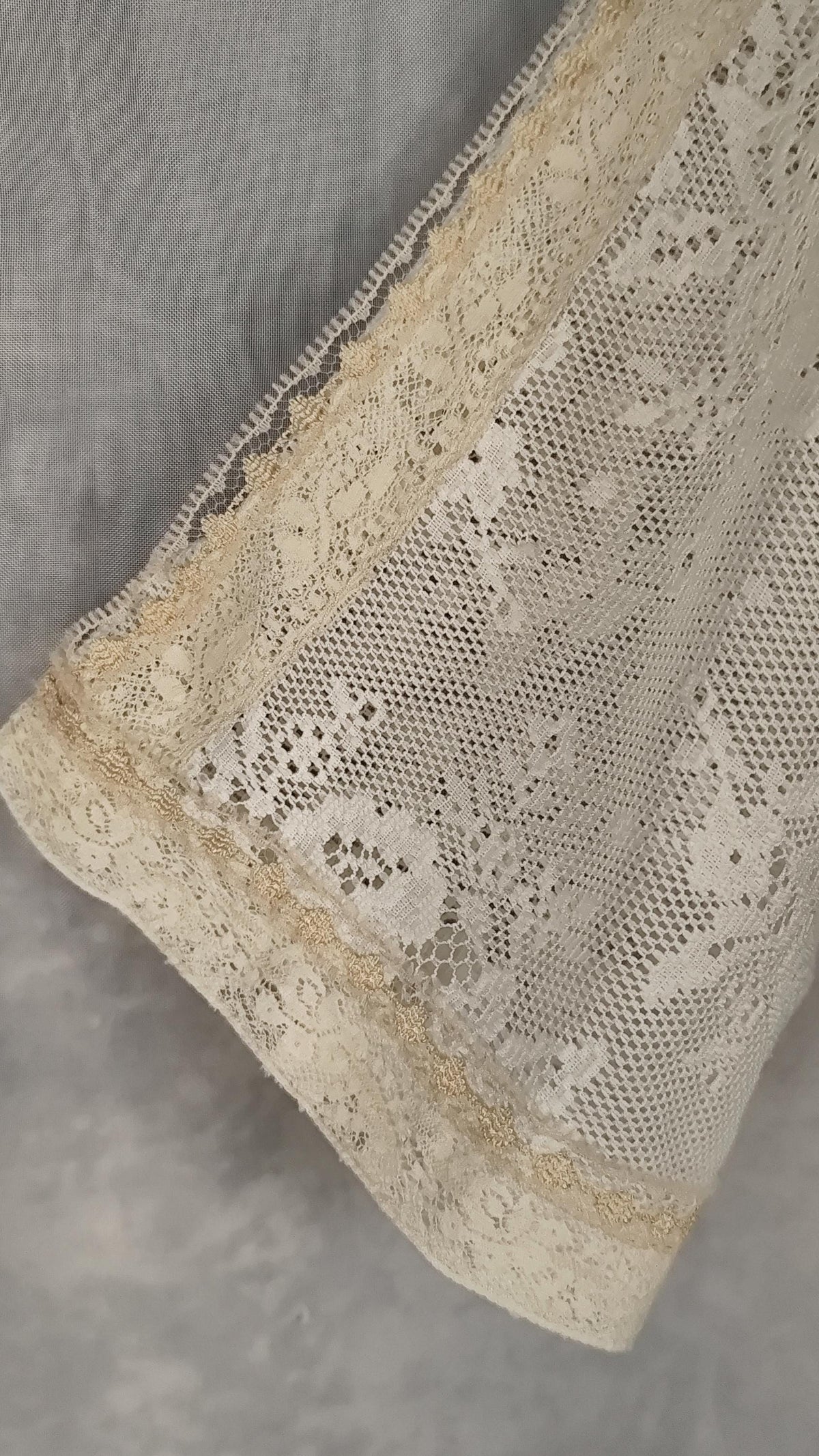 Close-up of a sheer lace top in pearl, showing floral lace patterns and trim, draped over a black hanger against a soft gray background.