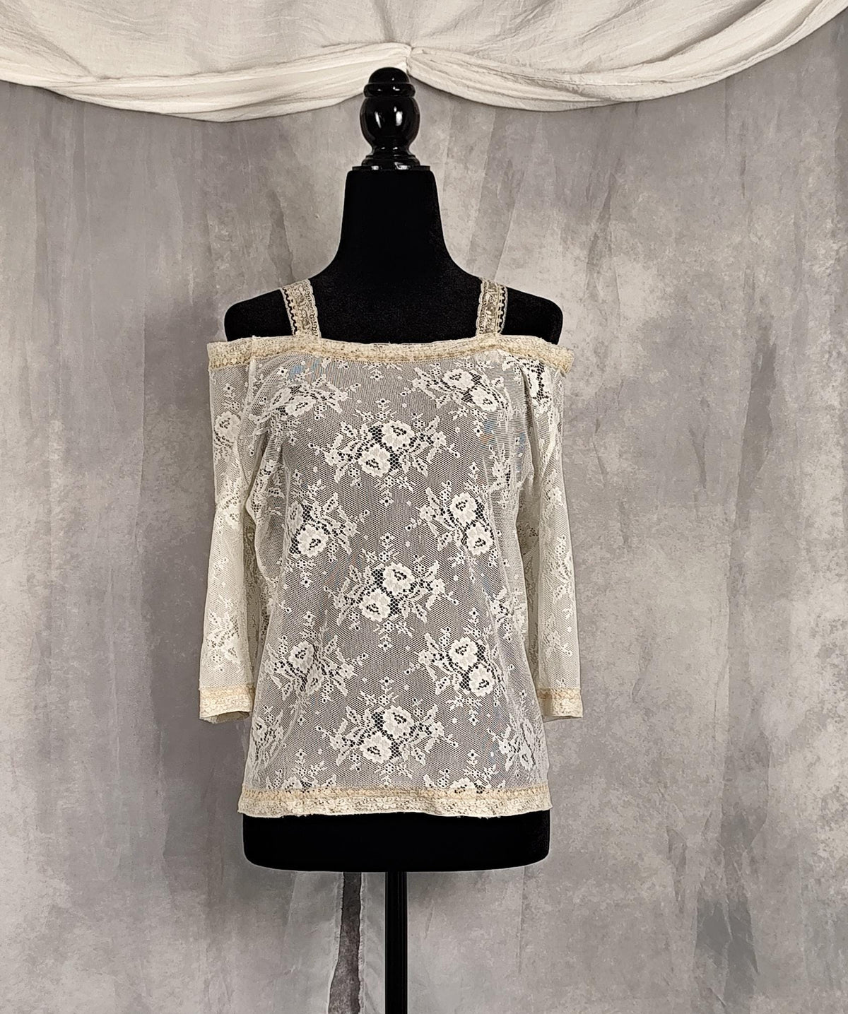 Front view of an off-the-shoulder lace top in cream and pearl, displayed on a black dress form mannequin against a textured gray backdrop with draped white fabric at the top.