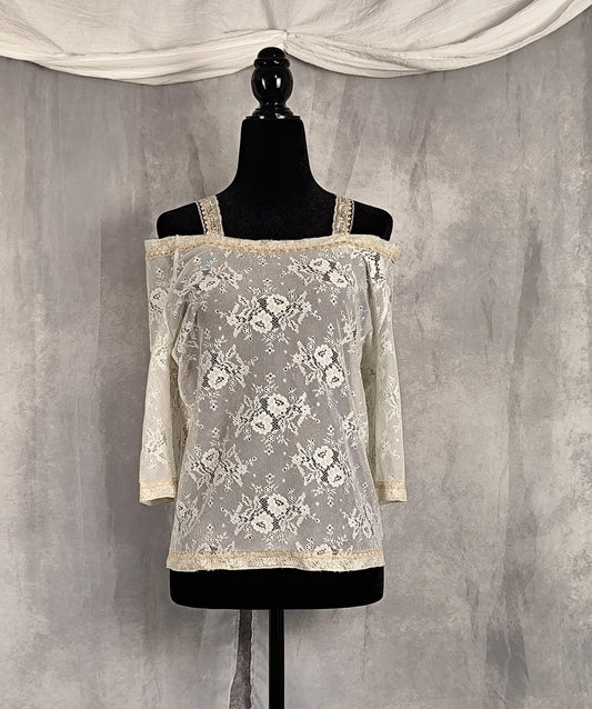 Front view of an off-the-shoulder lace top in cream and pearl, displayed on a black dress form mannequin against a textured gray backdrop with draped white fabric at the top.