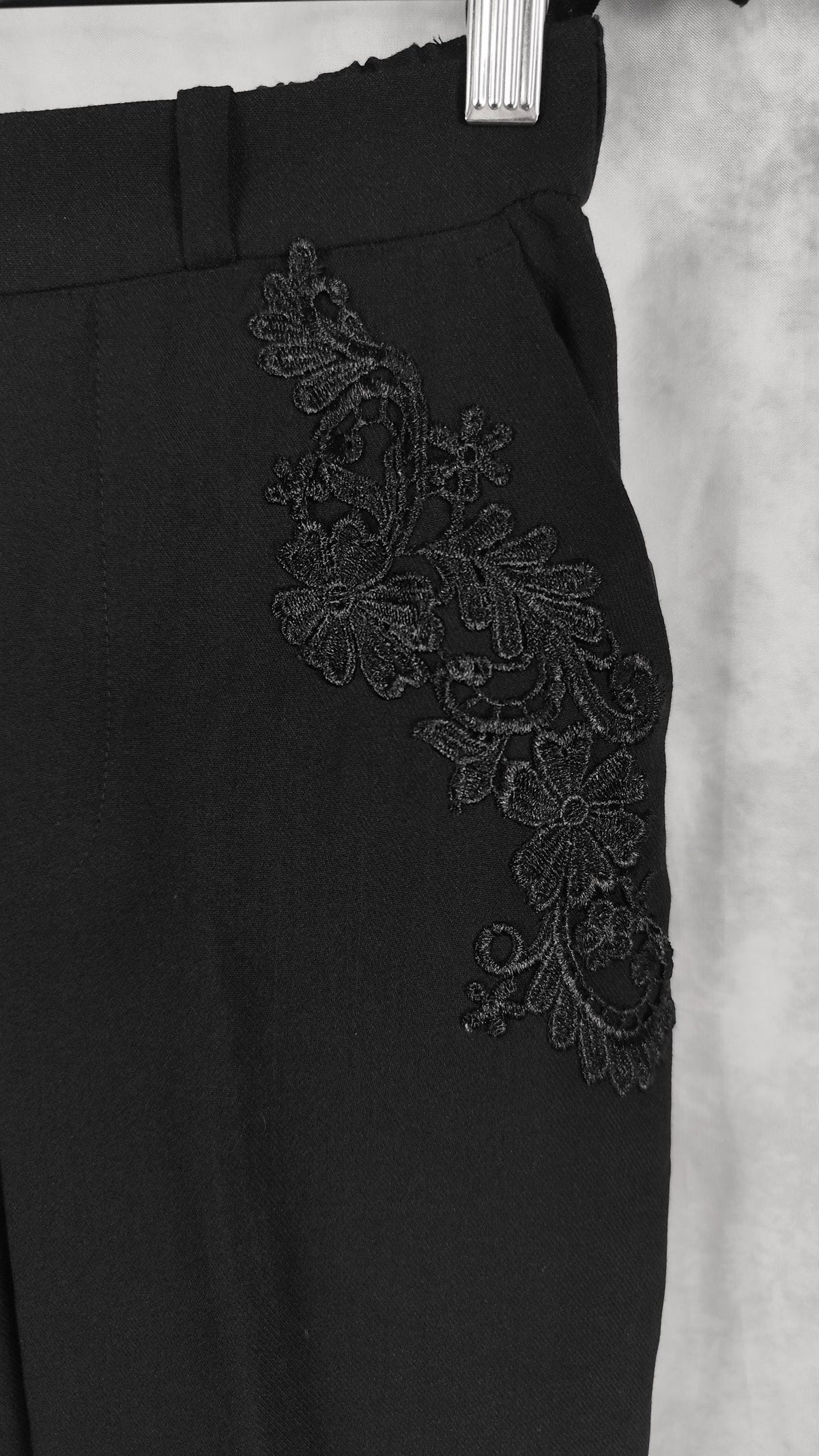 Black trousers featuring an intricate floral lace applique detail on the side, shown hanging against a textured gray background.
