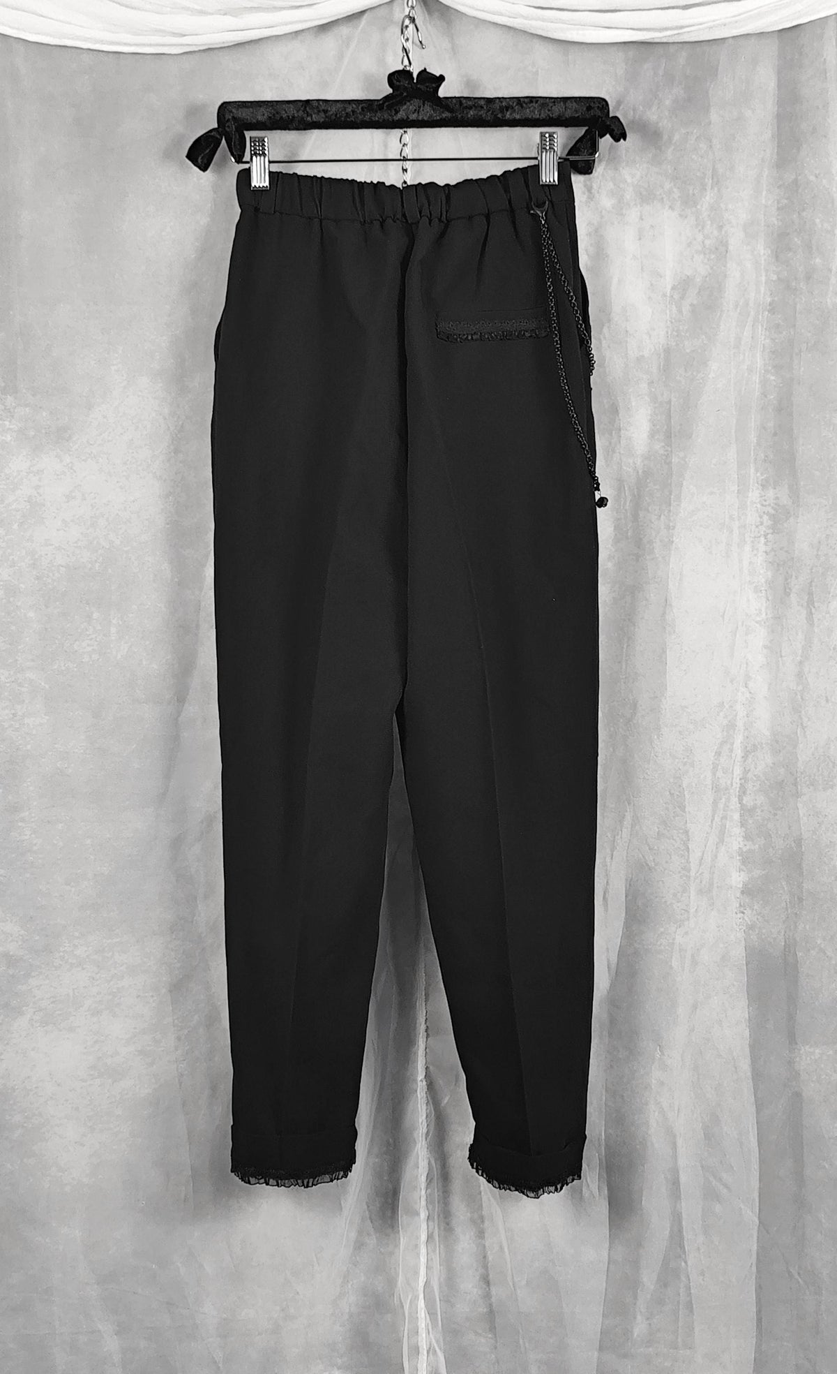 Back view of a black cropped pants with a drawstring waist and side pocket, hanging on a black hanger against a gray draped fabric backdrop.