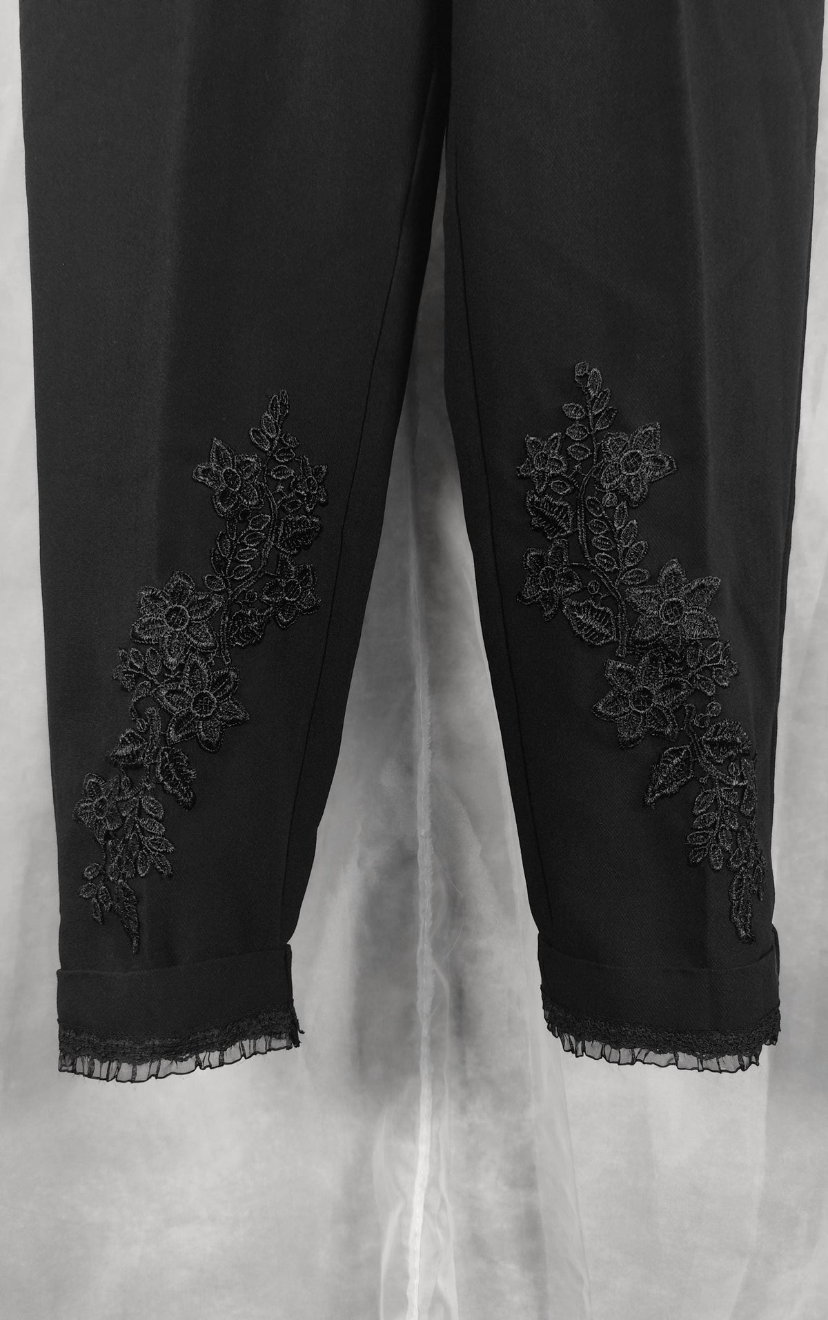 Black cropped trousers with floral lace applique detailing on the lower legs, shown against a light gray fabric backdrop.