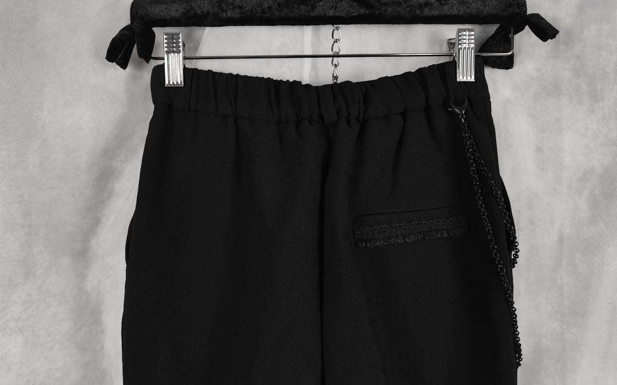 Black crop trousers with an elastic waistband, hanging from a black hanger with metal clips against a textured gray background.