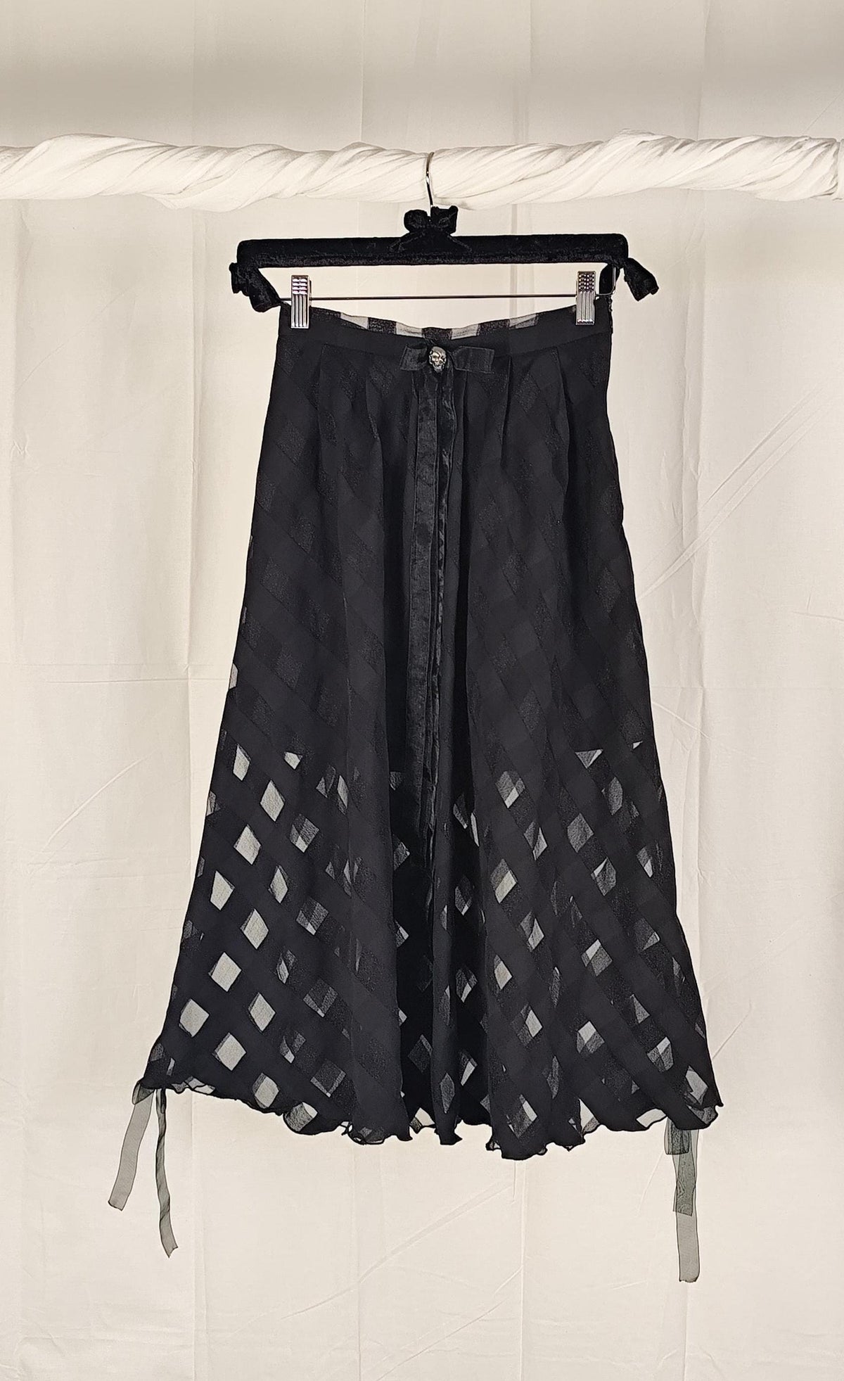 A black midi skirt with a diamond lattice cutout pattern, hanging on a black hanger against a white fabric backdrop.