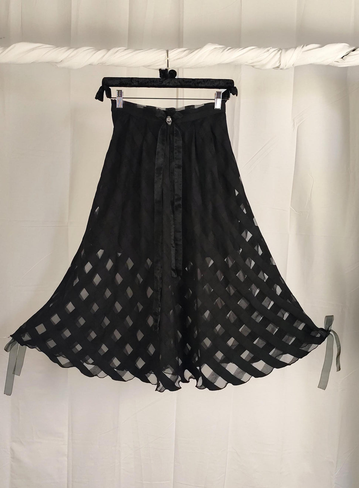 A black sheer mesh skirt with a checkered pattern and tie details, hanging on a black hanger against a white fabric backdrop.