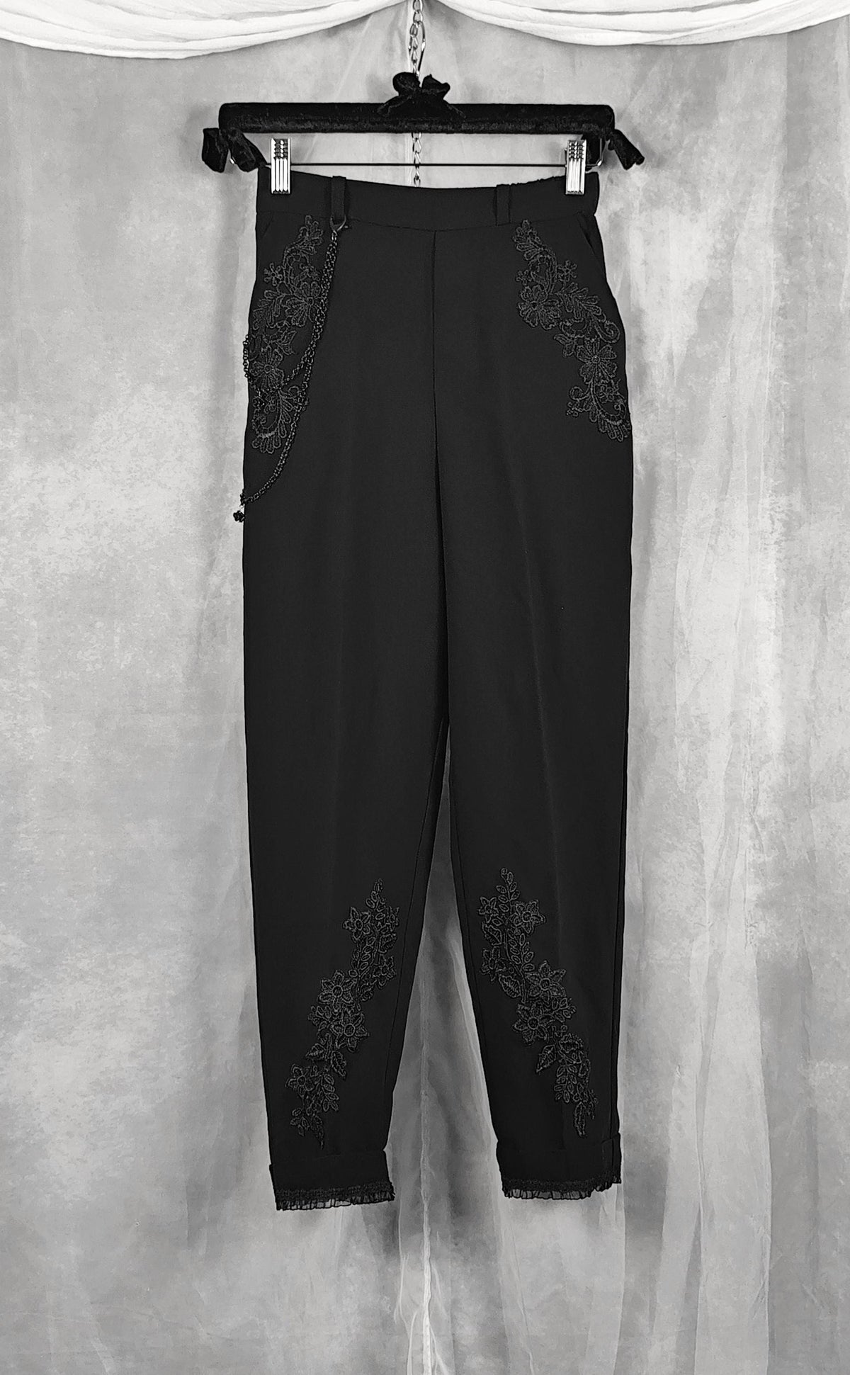 Front view of a black cropped trousers with intricate floral lace applique detailing on the sides and lower legs, hanging on a black hanger against a textured gray backdrop.