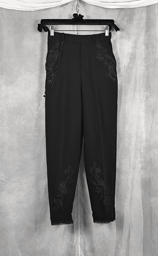 Front view of a black cropped trousers with intricate floral lace applique detailing on the sides and lower legs, hanging on a black hanger against a textured gray backdrop.