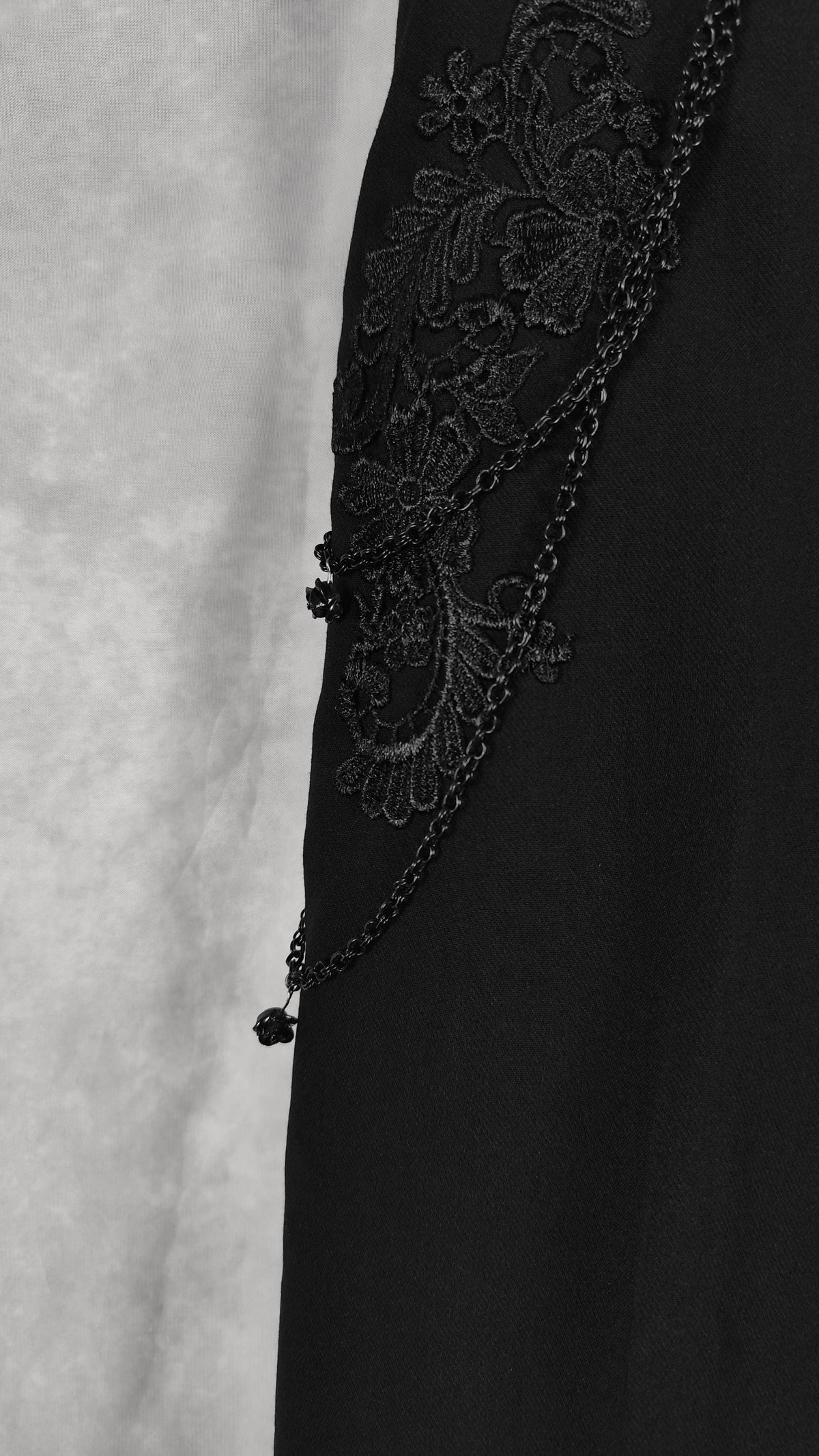 Close-up of a black garment featuring intricate black floral lace embroidery and a beaded chain detail against a light gray textured background.