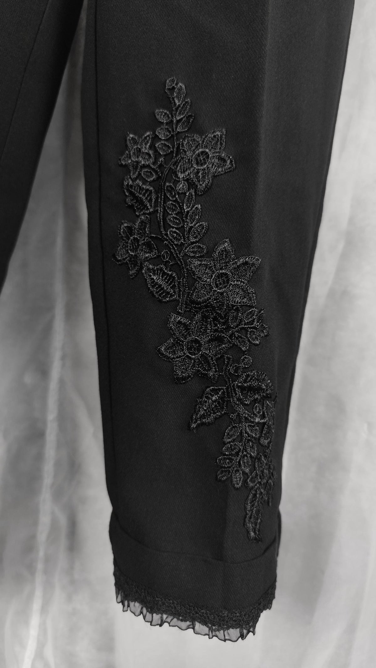 Black crop pants with a vertical floral lace applique detail on the lower leg and a lace hem, shown against a light draped fabric background.