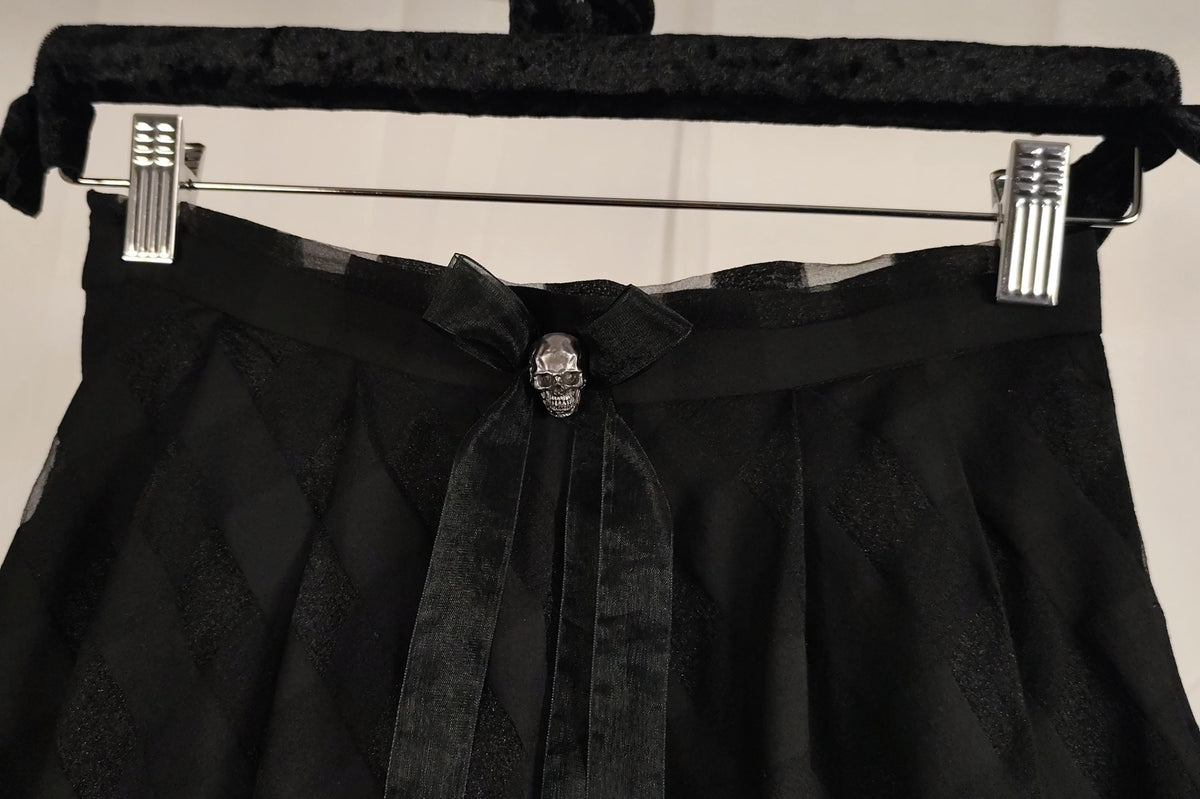 Black pleated skirt with a wide waistband, sheer side panels, and a bow featuring a silver metal skull, hanging on a black velvet hanger with silver metal clips.