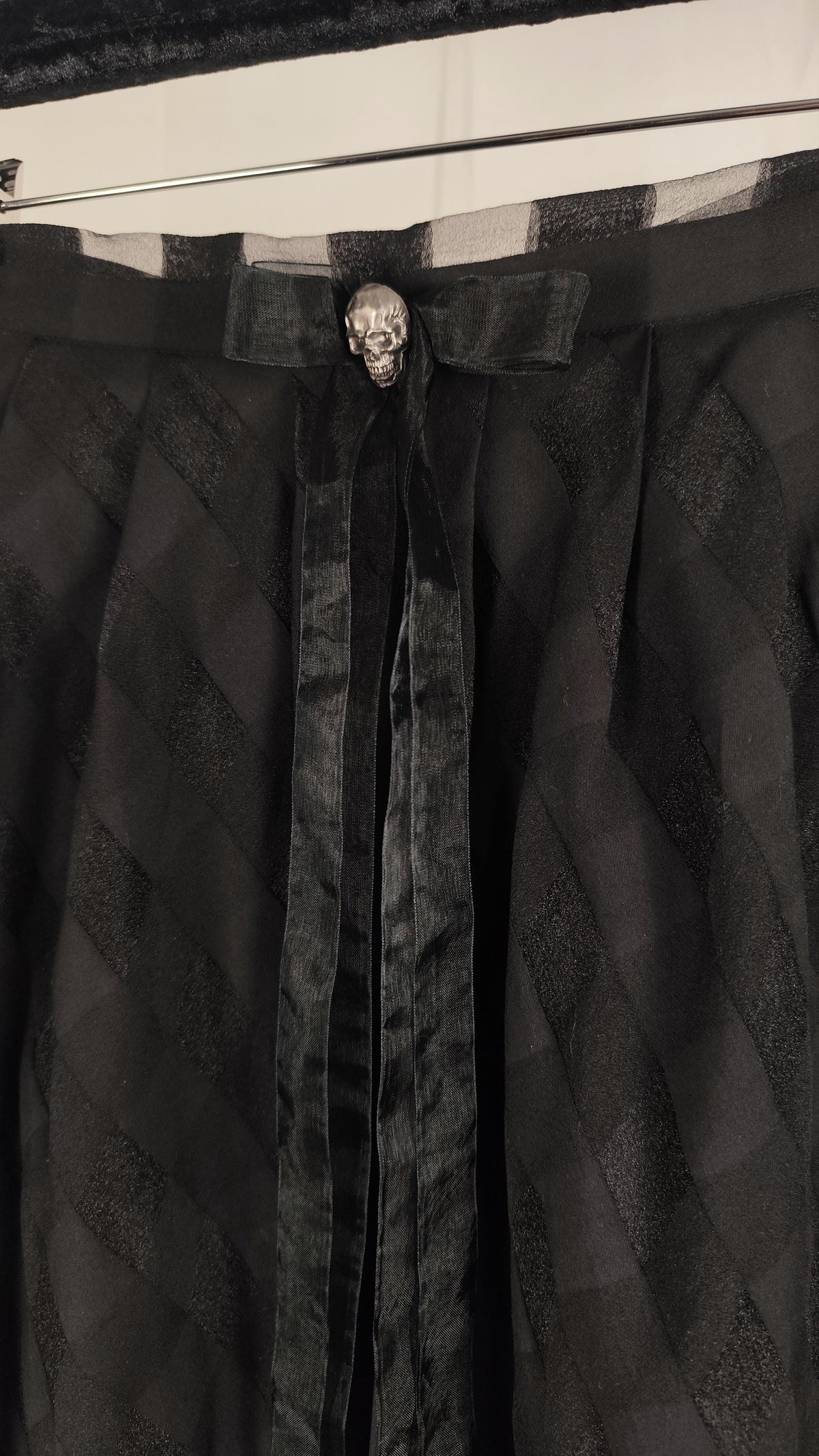 A close-up of a black gothic-style skirt featuring a textured diamond-patterned fabric, a central black velvet ribbon, and a silver skull brooch at the waist.