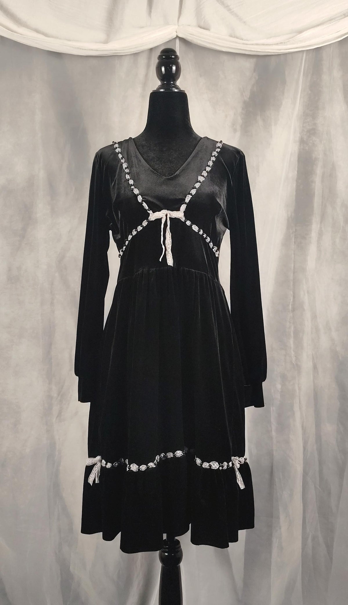 Front view of a black velvet long-sleeve dress with white beaded trim and tie details, displayed on a black mannequin against a draped gray fabric backdrop.