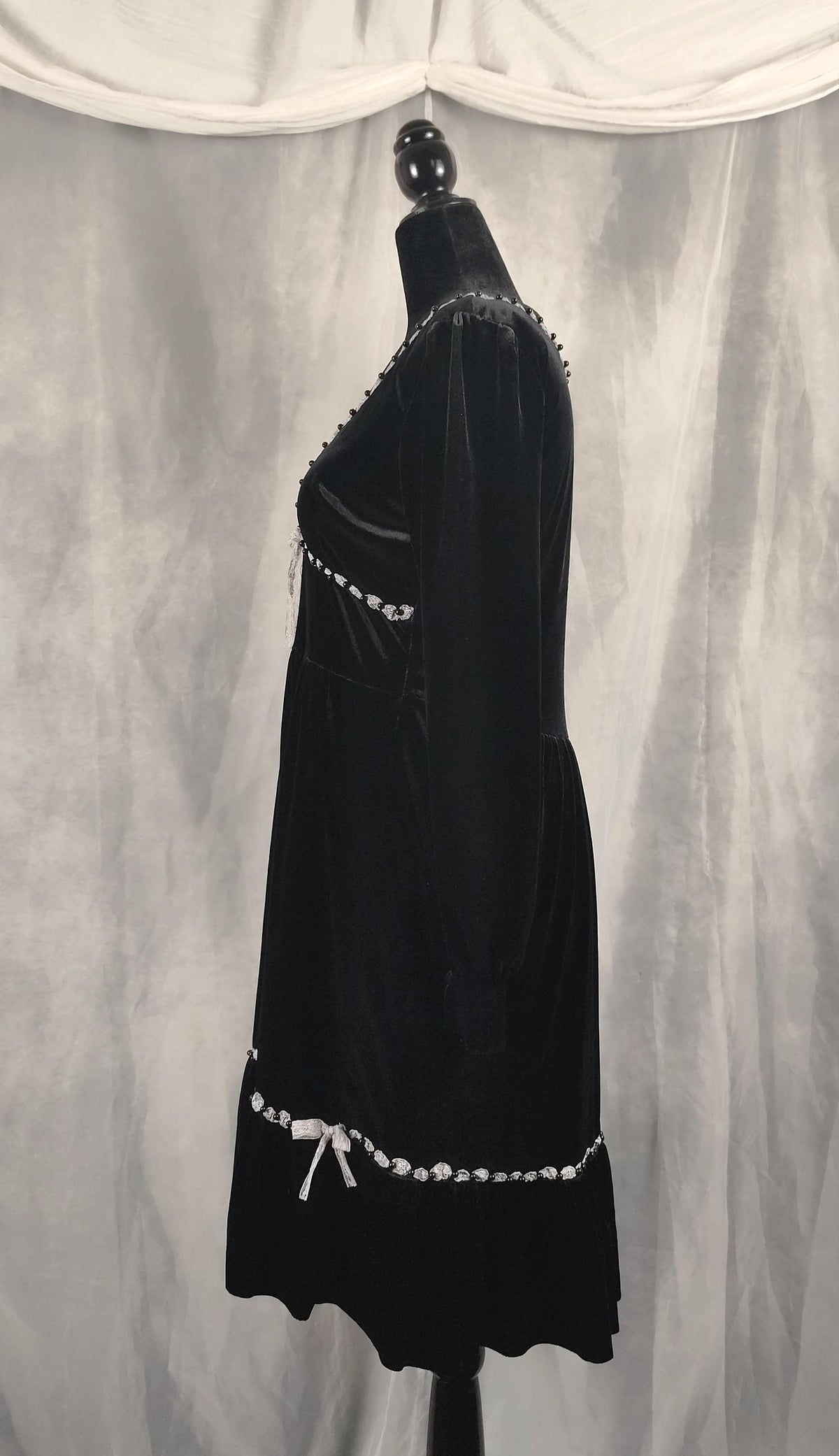 Side view of a black velvet dress displayed on a black mannequin against a draped gray fabric backdrop. The dress features white beaded trim along the neckline, waist, and hem, and a white bow detail at the bottom.
