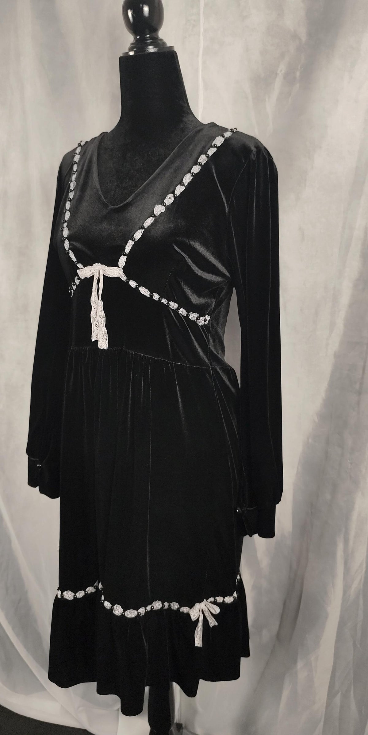 A black velvet long-sleeve A-line dress with white beaded trim and white tie details, displayed on a black mannequin against a draped white fabric backdrop.