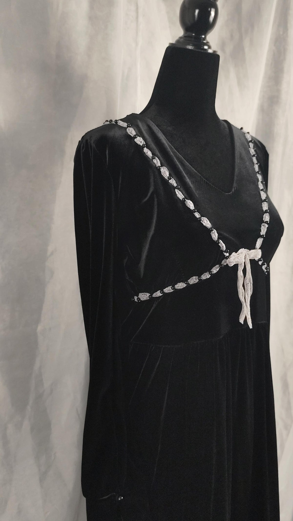 A black velvet dress with black beads trim and a white bow, displayed on a black mannequin torso against a draped gray fabric backdrop.