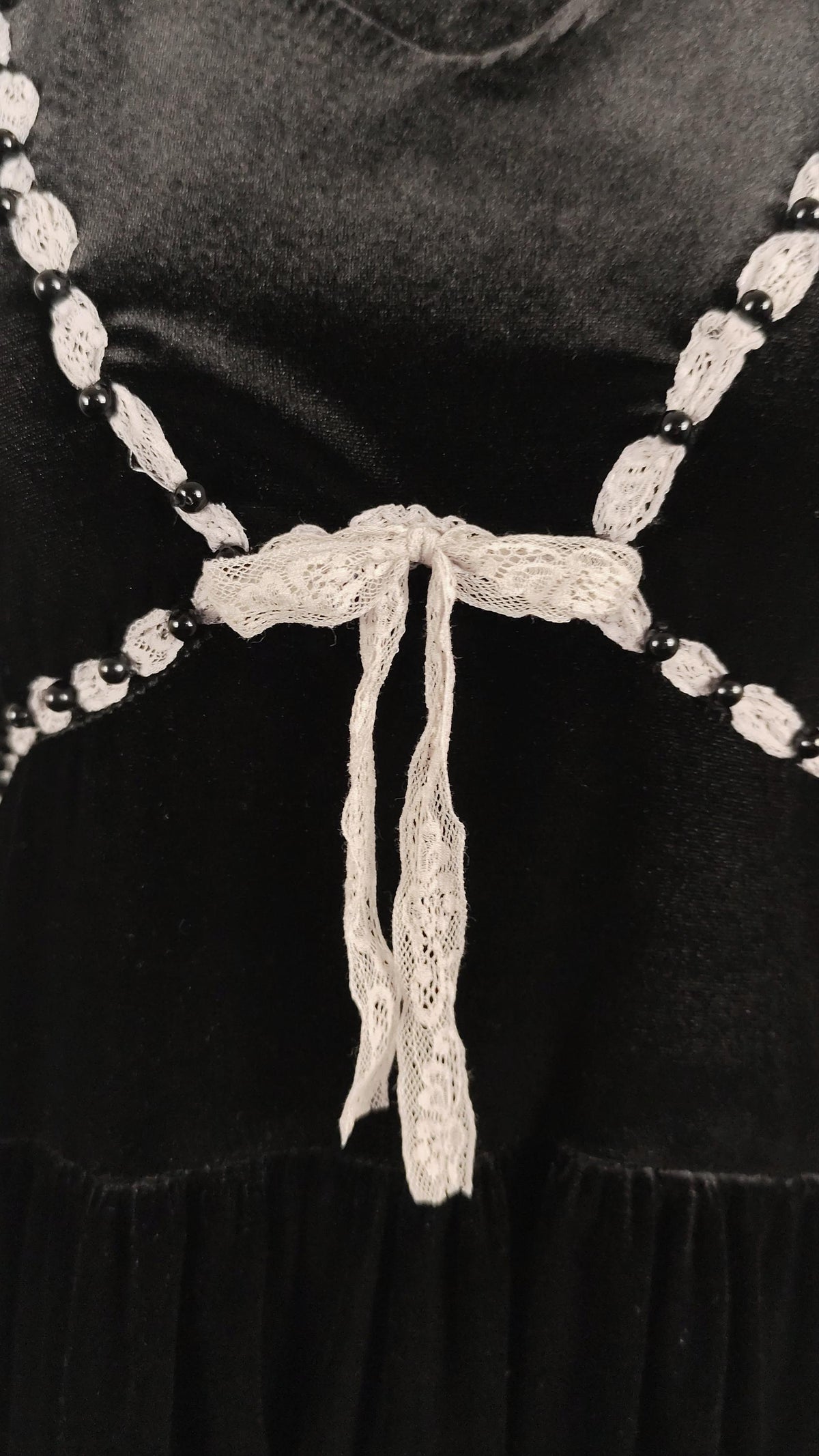 A close-up of a black dress featuring a decorative off-white lace bow at the center of the waist, connected by a chain embellished with black beads.