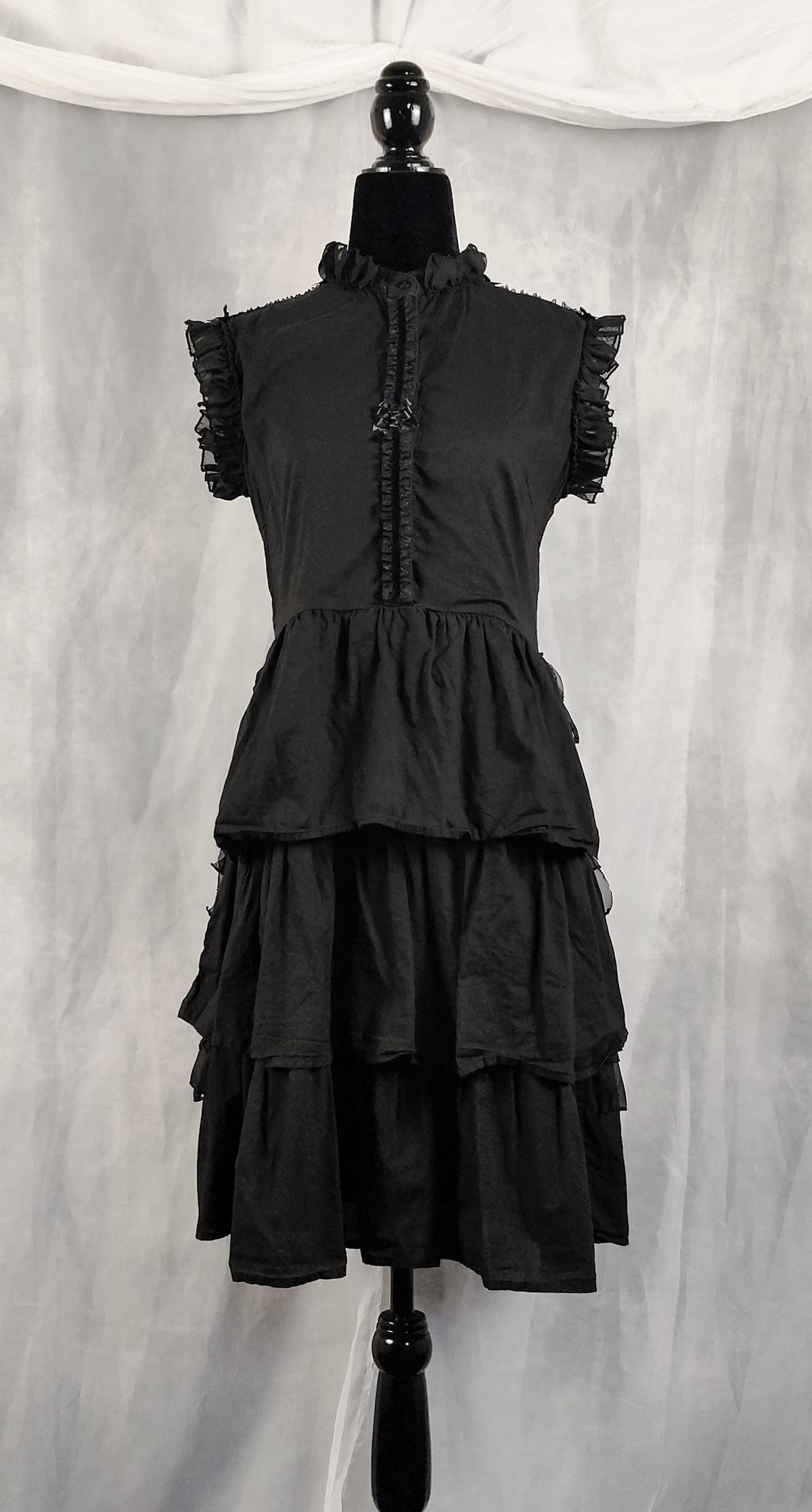 Front view of a black tiered ruffled dress displayed on a black mannequin against a gray draped fabric backdrop.