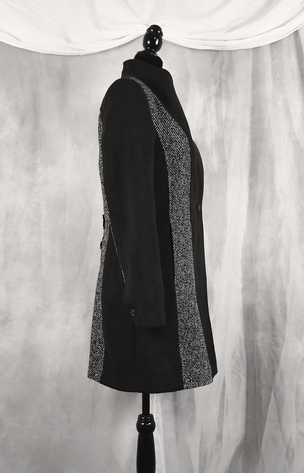 Side view of black coat with textured gray and white patterned side panels, displayed on a black dress form mannequin against a draped gray fabric backdrop.