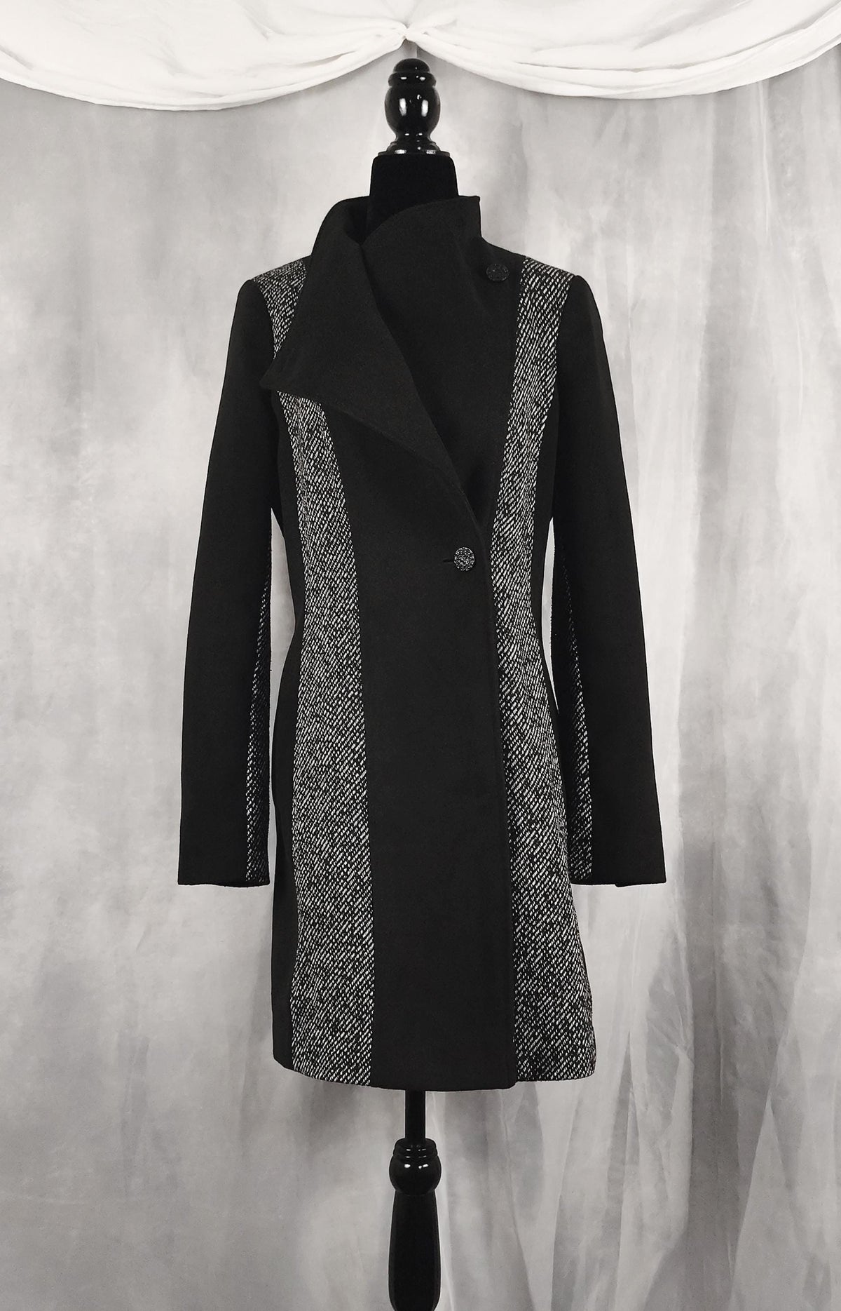 Front view of a black longline coat with textured gray and white patterned side panels, displayed on a black dress form mannequin against a draped gray fabric backdrop.