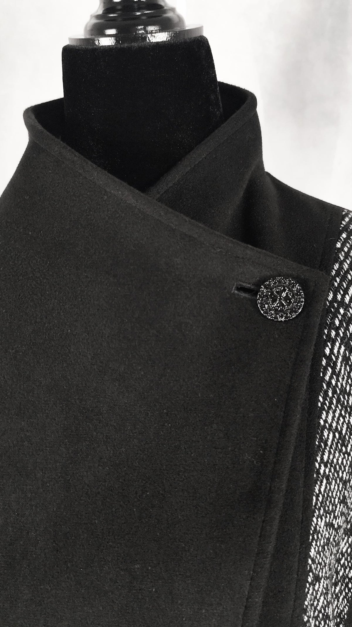 Close-up of a black wrap-style coat displayed on a black mannequin torso, with a decorative round button and a textured patterned side panel visible against a light gray background.