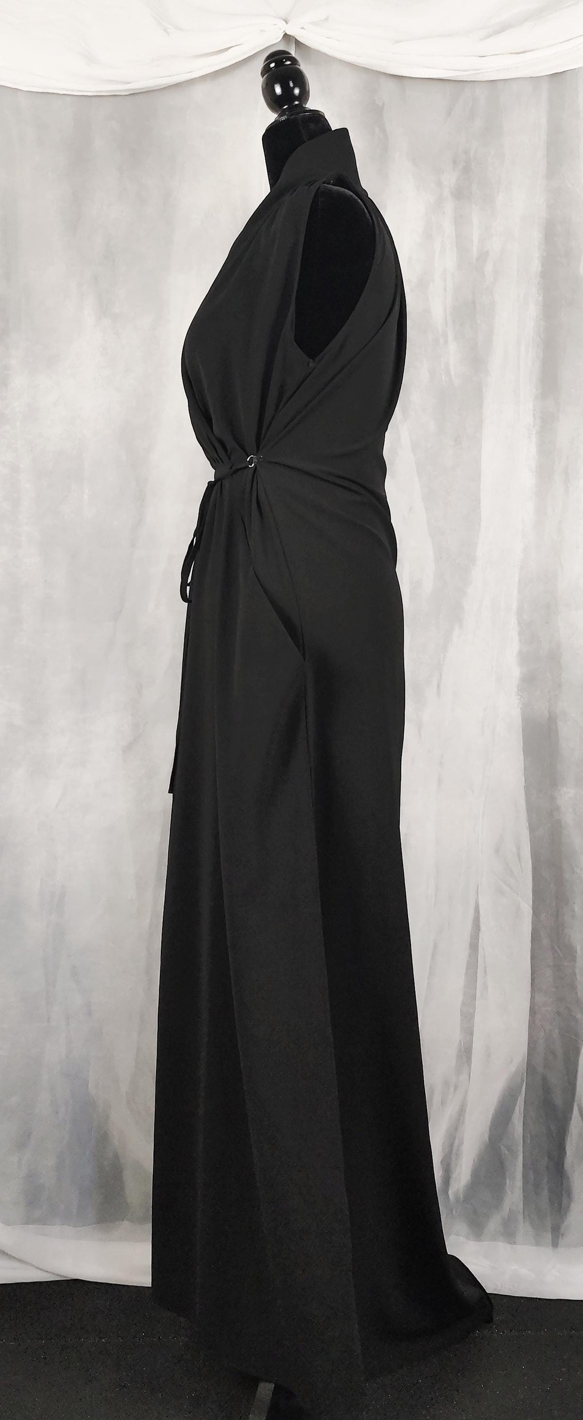 Side view of a  sleeveless black maxi dress displayed on a mannequin. The dress features a crossover V-neckline, an adjustable drawstring waist tie, and a long, flowing skirt with side pockets