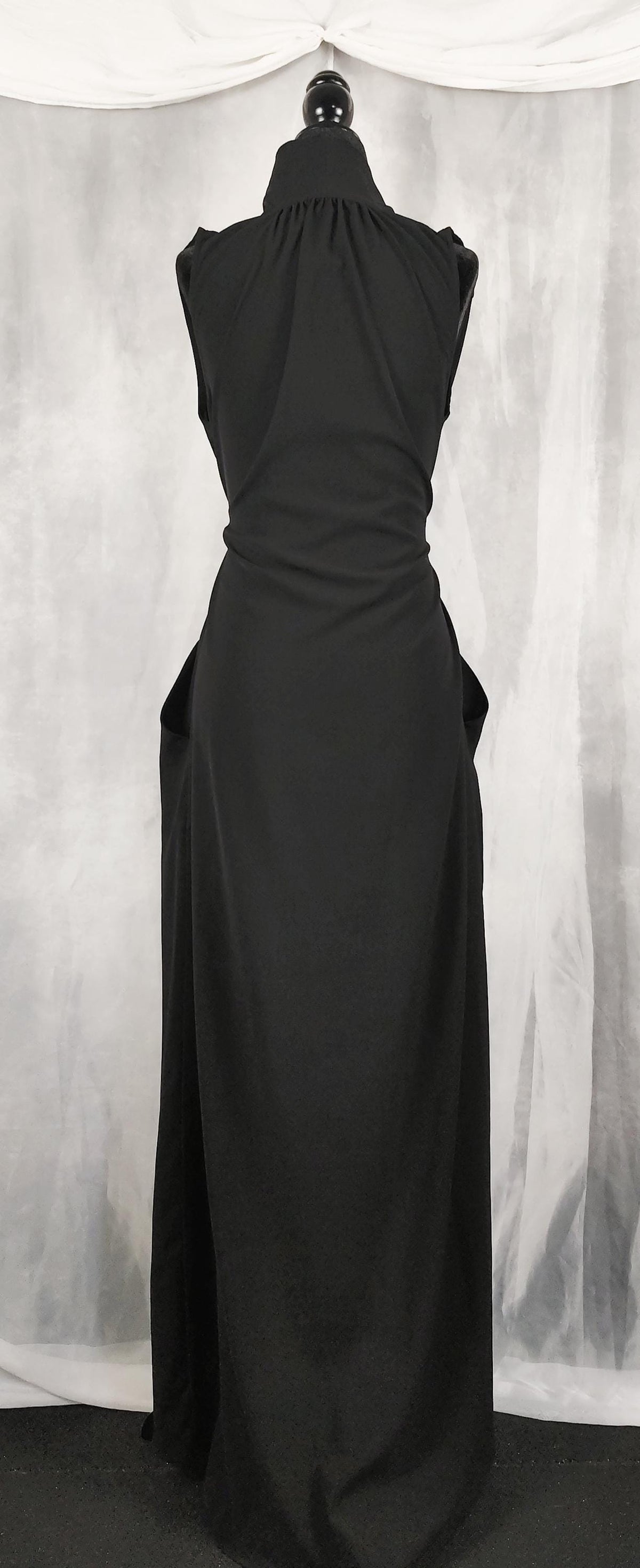 Back view of a sleeveless black maxi dress displayed on a mannequin. The dress features a crossover V-neckline, an adjustable drawstring waist tie, and a long, flowing skirt with side pockets