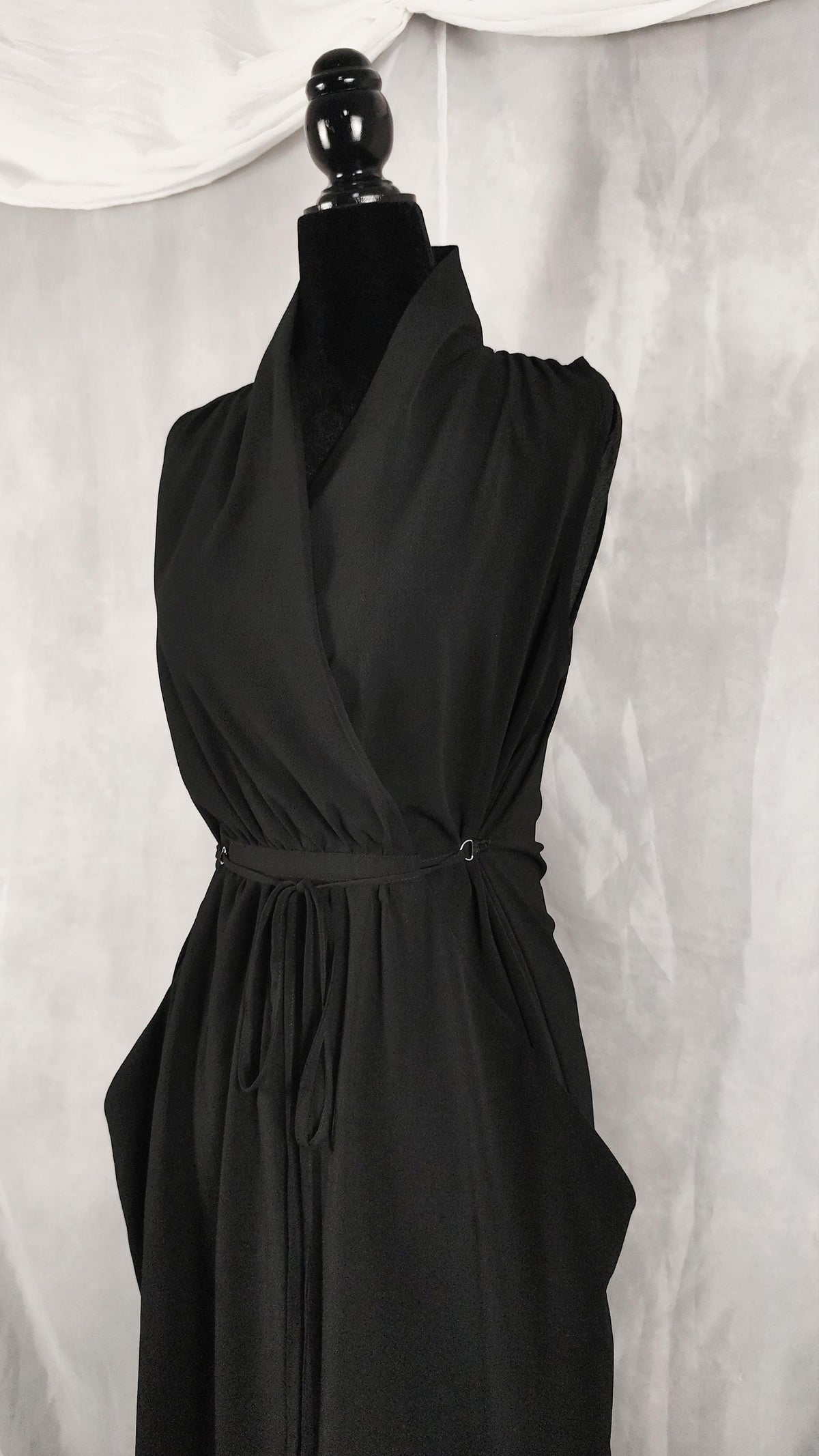 A black sleeveless wrap-style dress displayed on a black mannequin against a light gray draped fabric backdrop.