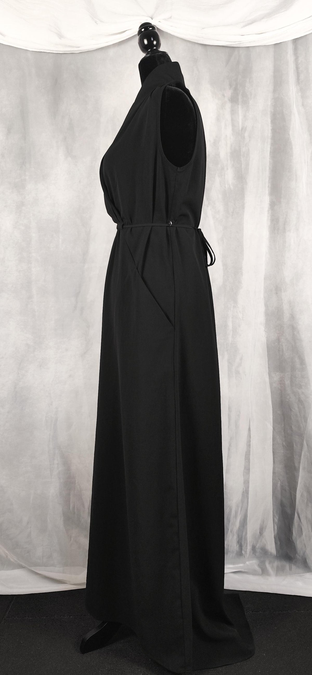 Side view of a sleeveless black maxi dress displayed on a black dress form mannequin against a gray draped fabric backdrop.