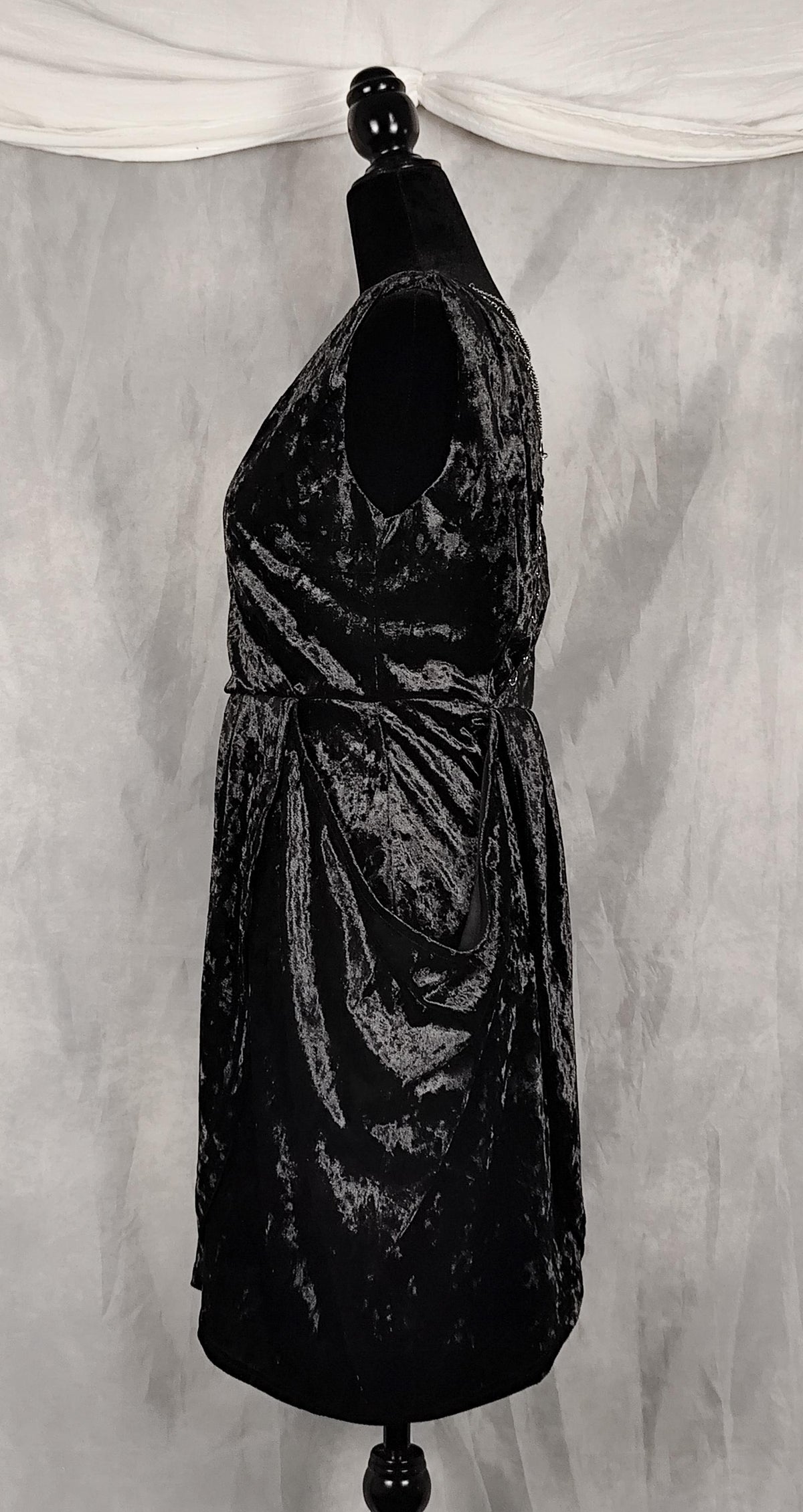 A sleeveless black crushed velvet dress displayed on a black mannequin torso against a gray draped fabric backdrop.