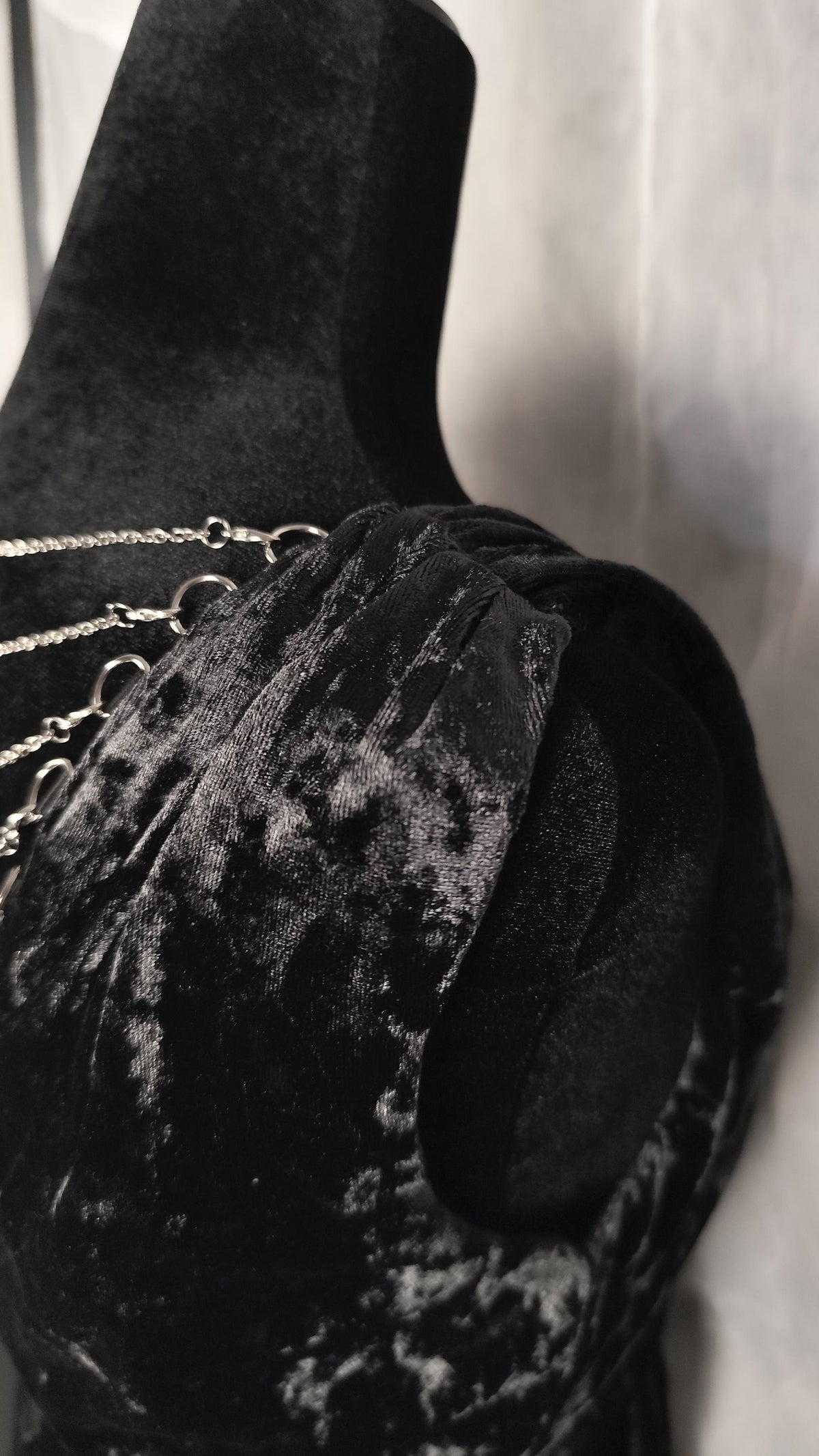 A close-up of a black velvet dress featuring a textured crushed velvet finish and delicate silver chain straps, displayed on a mannequin against a light gray background.