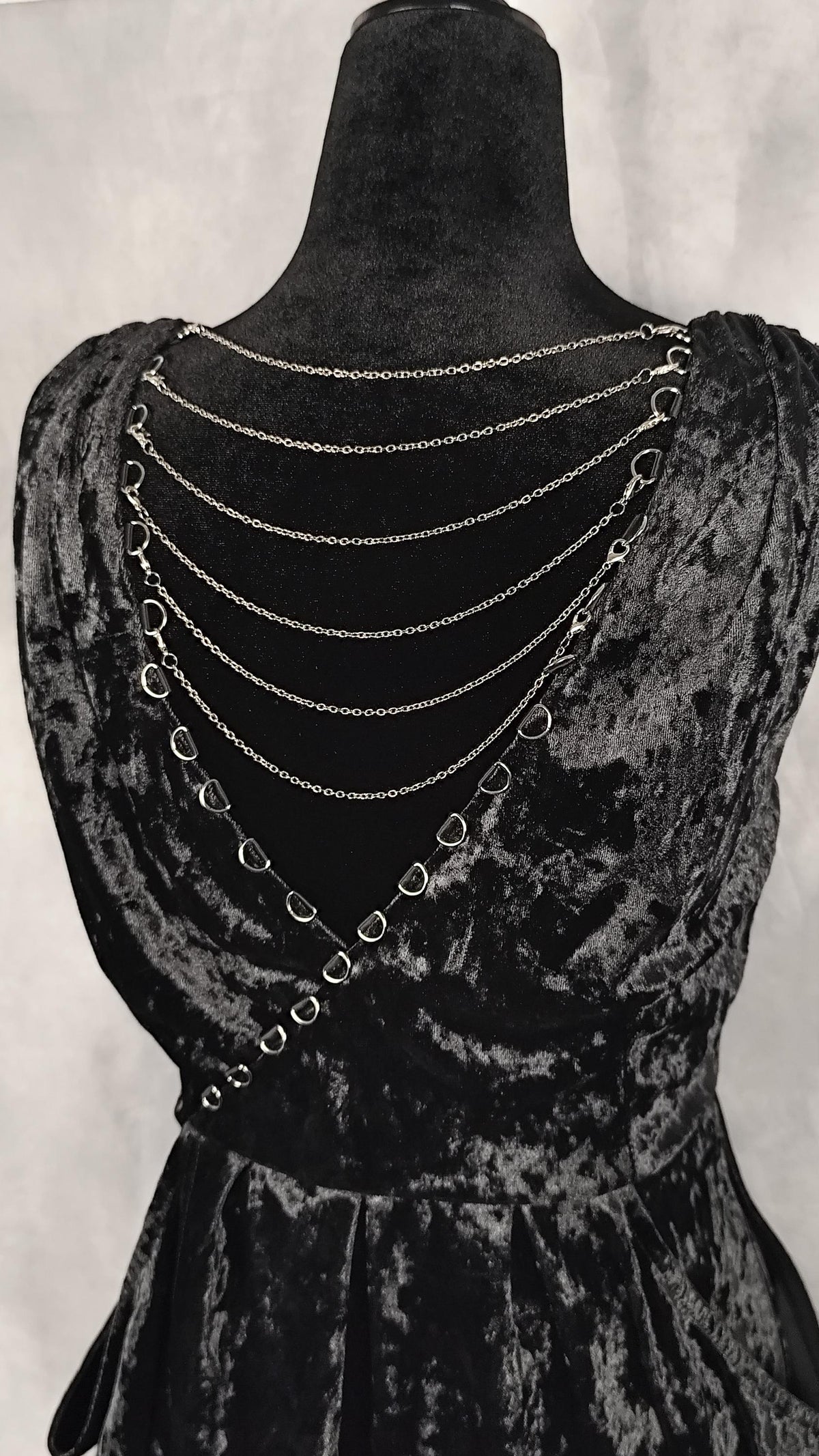 A black velvet dress displayed on a black mannequin, featuring a dramatic back design with multiple draped silver chains and interlocking ring details.