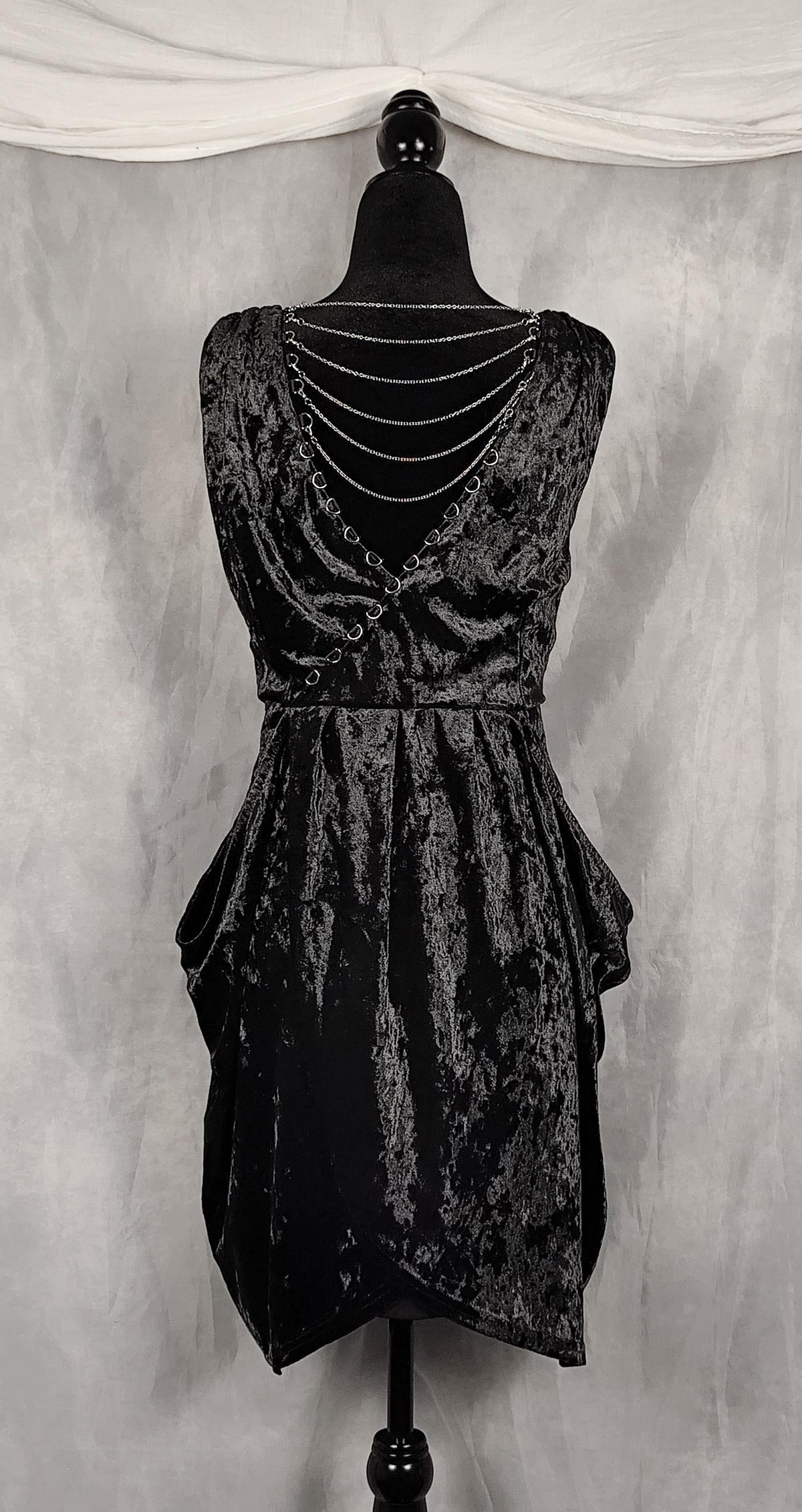A sleeveless black crushed velvet dress with a tiered ruffled skirt and a double-row of silver chain embellishments across the back, displayed on a black mannequin against a gray backdrop.