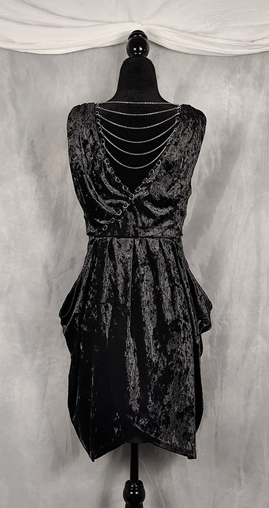 A sleeveless black crushed velvet dress with a tiered ruffled skirt and a double-row of silver chain embellishments across the back, displayed on a black mannequin against a gray backdrop.