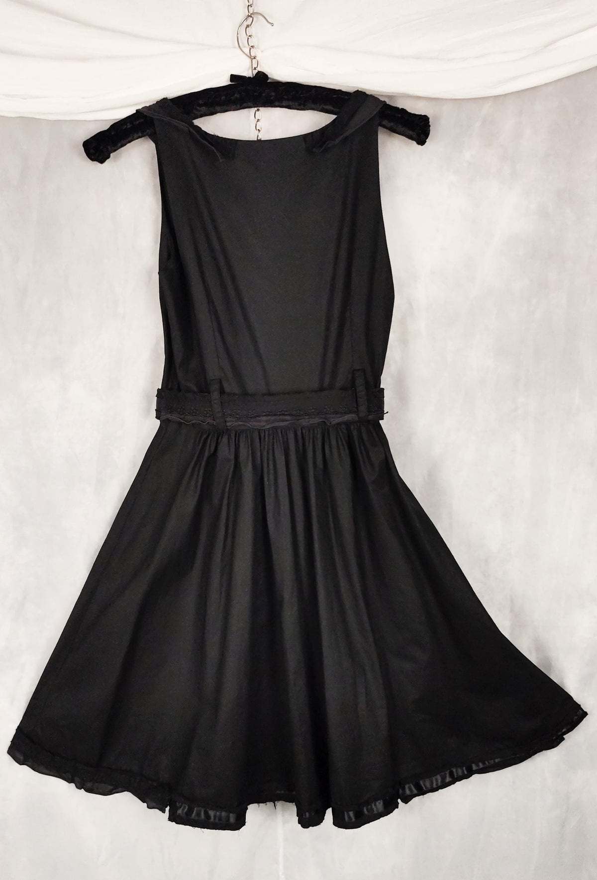 Back view of a black sleeveless dress with a structured corset-style bodice, and matching belt, hanging on a black hanger against a textured gray backdrop.