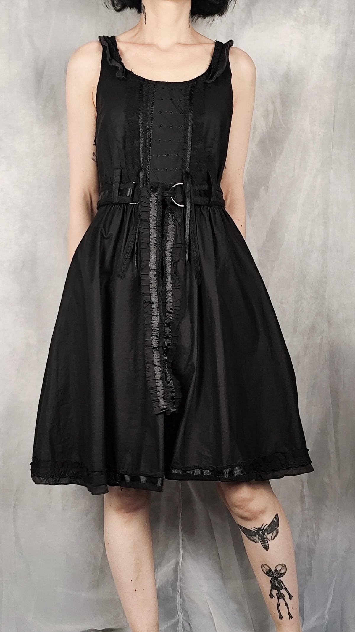 A black sleeveless A-line dress with ruffled trim, a gathered waist, and a central tie detail, shown on a model against a textured gray backdrop.