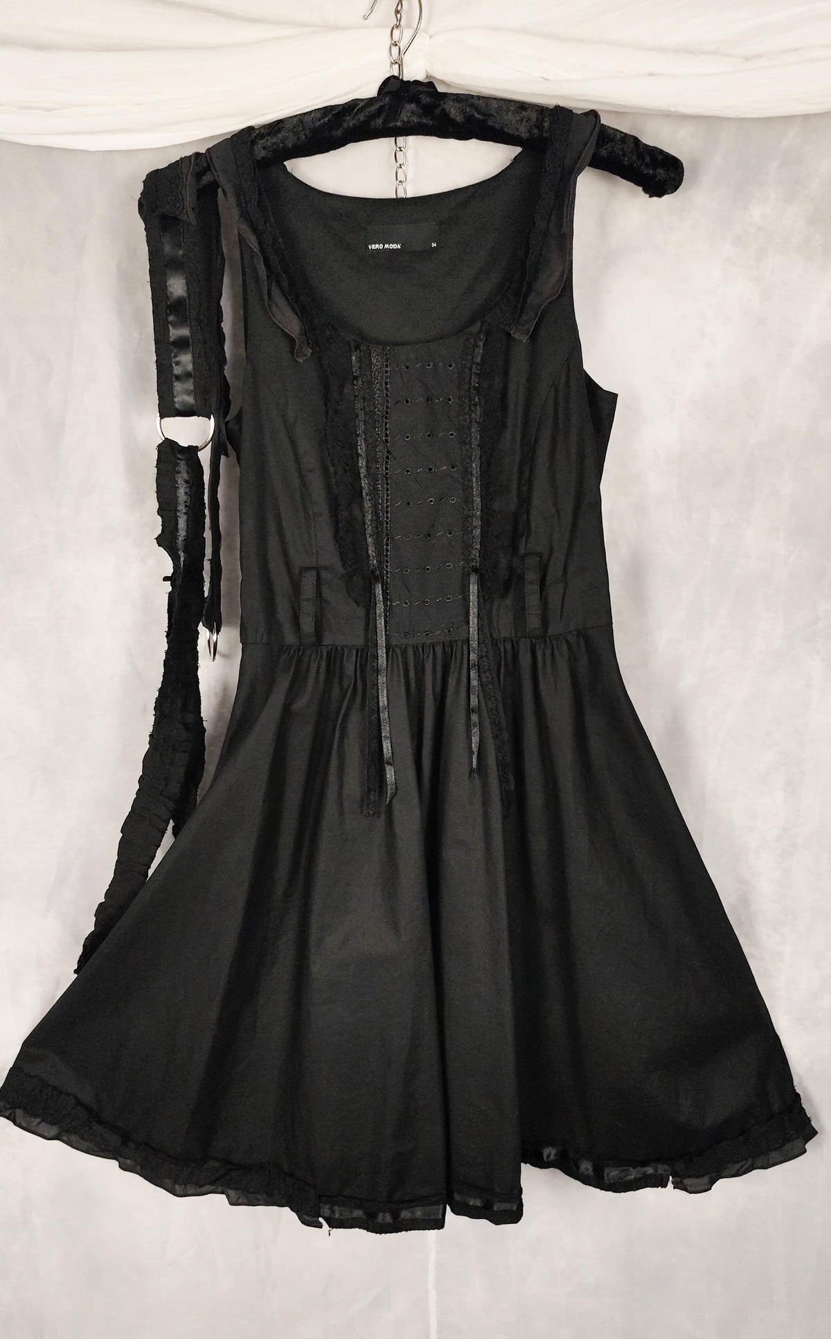 A black sleeveless dress with a gathered waist and sheer ruffled trim, hanging on a black hanger against a white fabric backdrop.