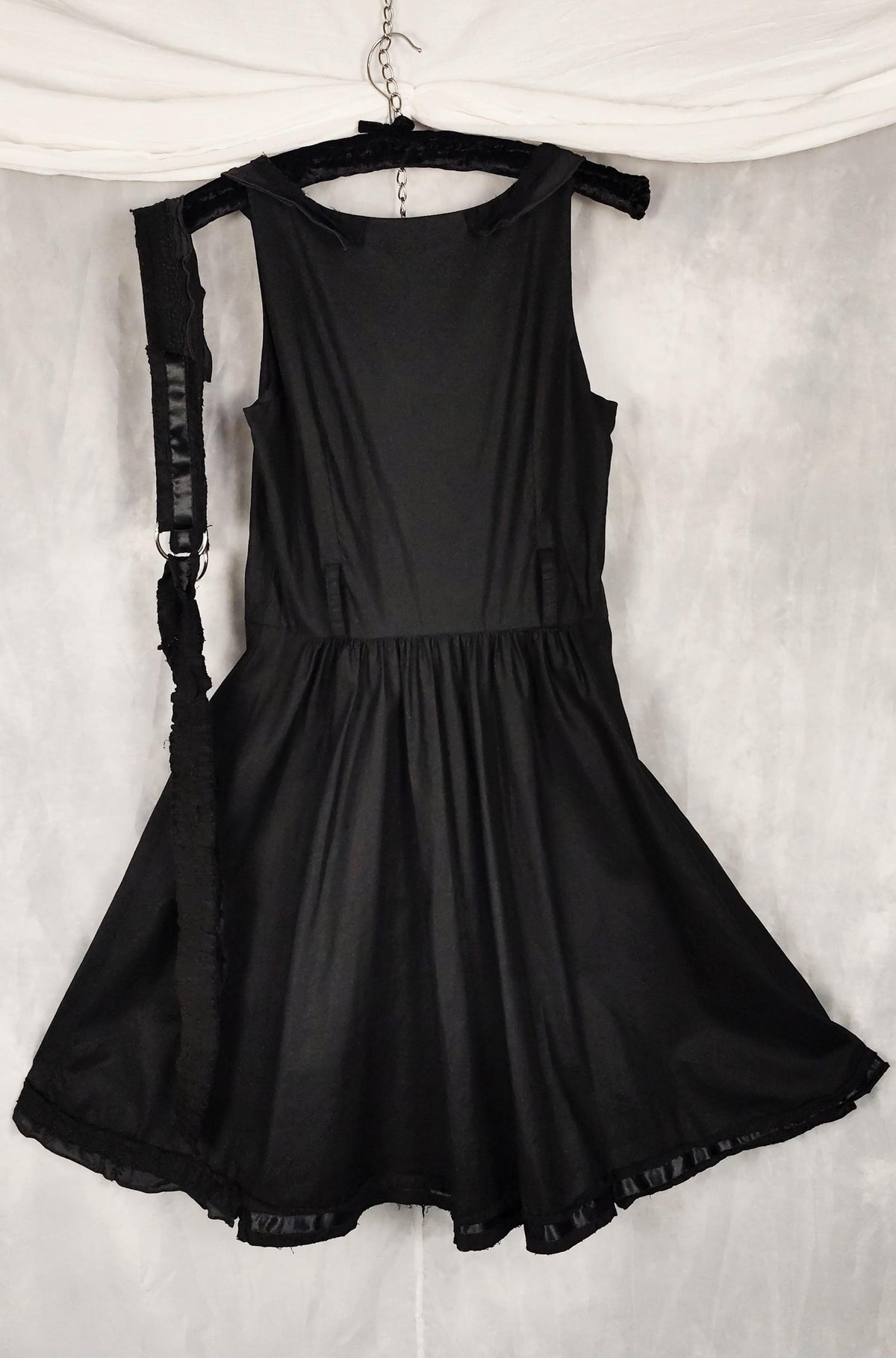 A black sleeveless dress with a gathered waist and flared skirt, hanging on a black hanger against a white draped backdrop.