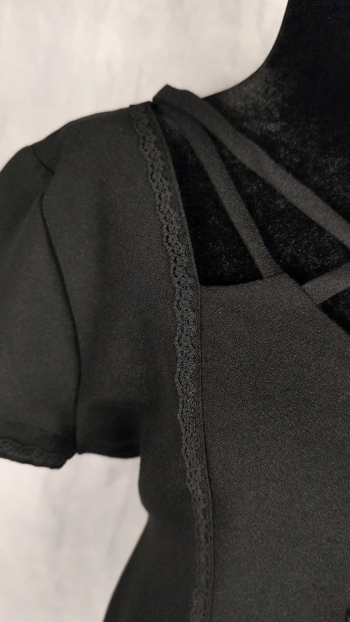 Close-up of a black short-sleeve midi dress on a mannequin, showing a textured front panel, a lace trim running down the center, and thin shoulder straps against a light gray background.