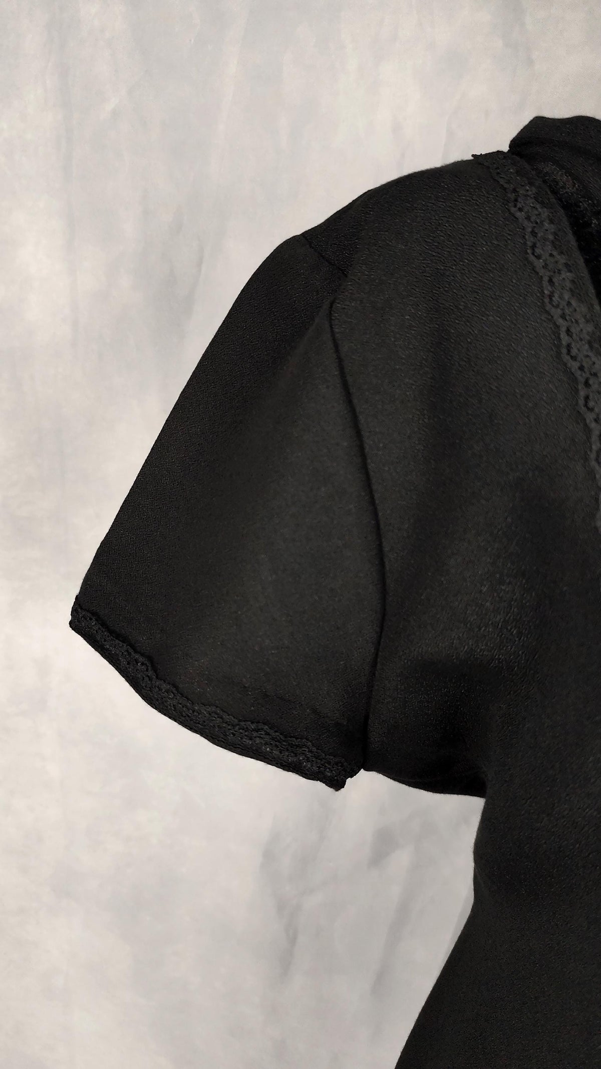 Close-up of a black short-sleeve midi dress with lace trim along the sleeve and neckline, shown against a light gray textured background.