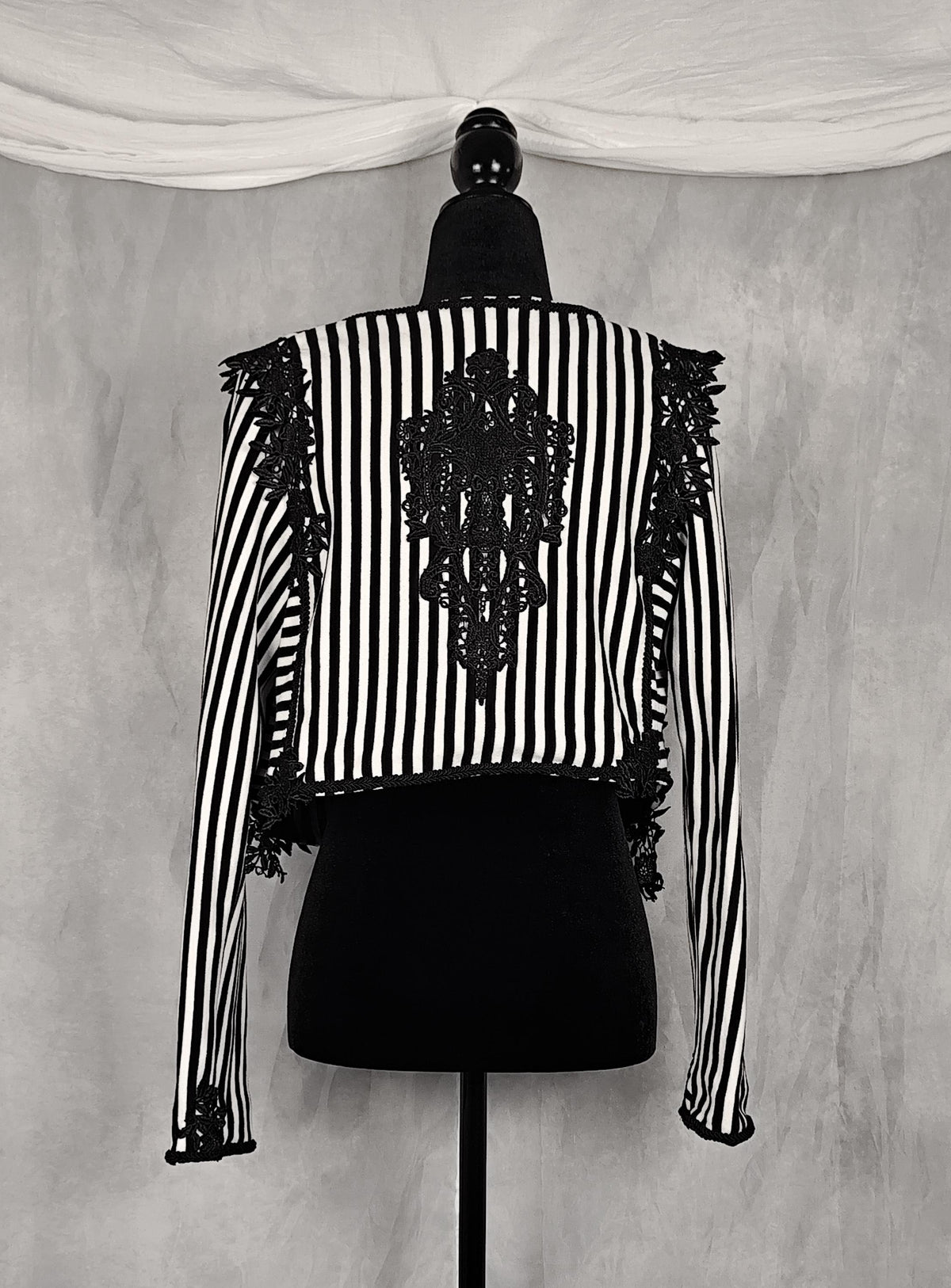 Back view of a black and white vertically striped long-sleeve jacket with black lace applique on the back and shoulders, displayed on a black dress form mannequin against a gray textured backdrop.