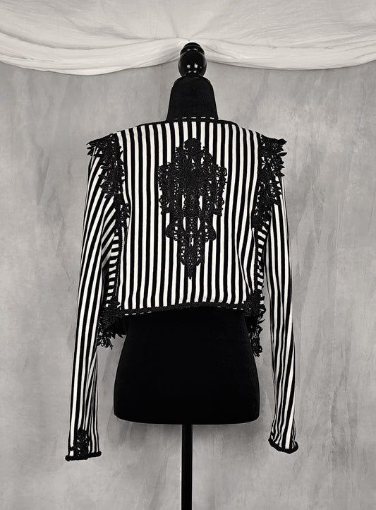 Back view of a black and white vertically striped long-sleeve jacket with black lace applique on the back and shoulders, displayed on a black dress form mannequin against a gray textured backdrop.