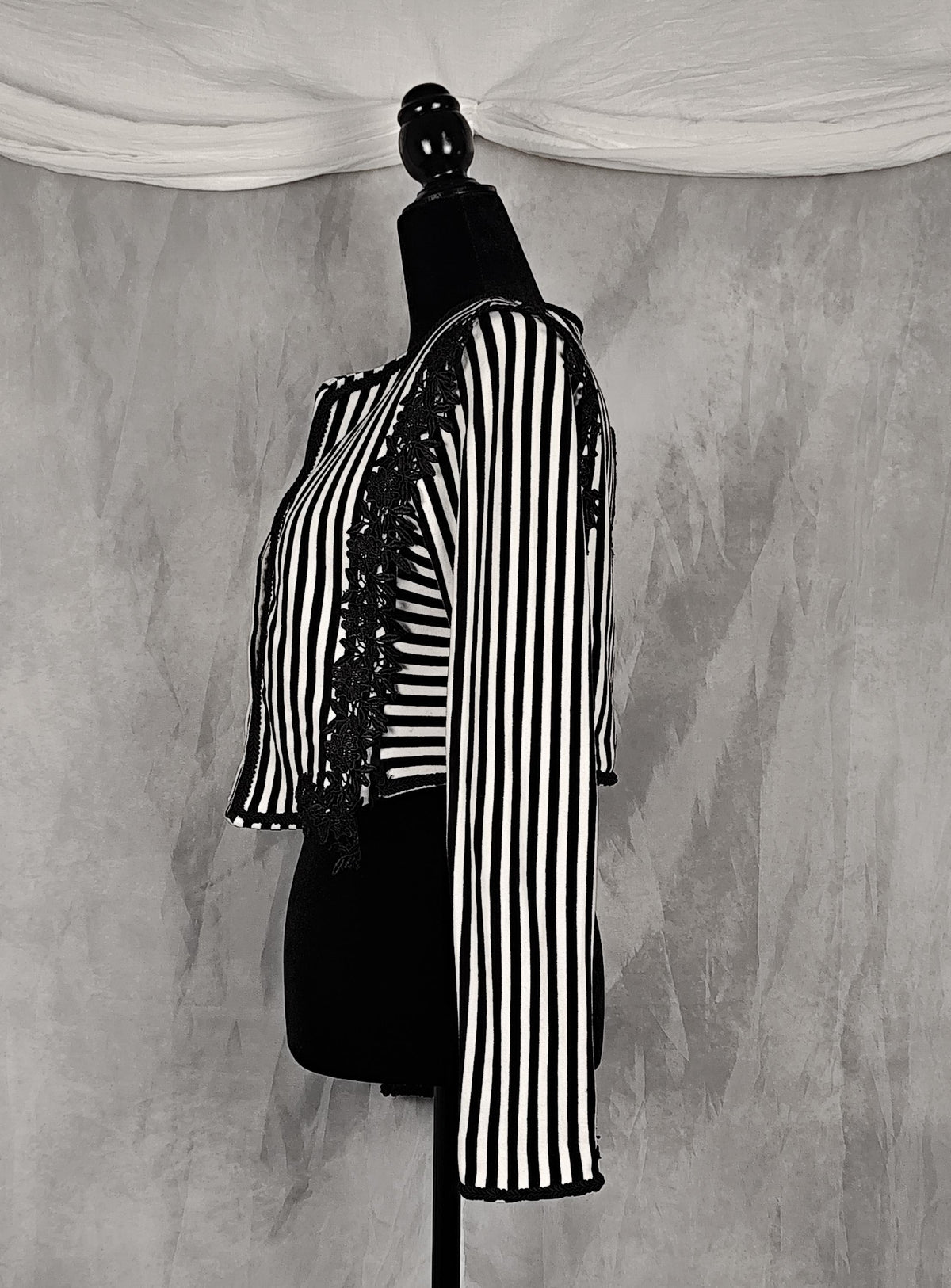 Side view of a black and white vertically striped long-sleeve blouse with black lace trim down the front, displayed on a black dress form mannequin against a textured gray backdrop.