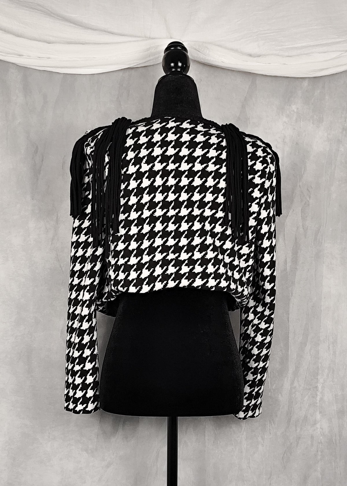 Back view of a black and white houndstooth cropped shrug jacket with shoulder cutouts and tassel ties, displayed on a black mannequin against a textured gray backdrop.