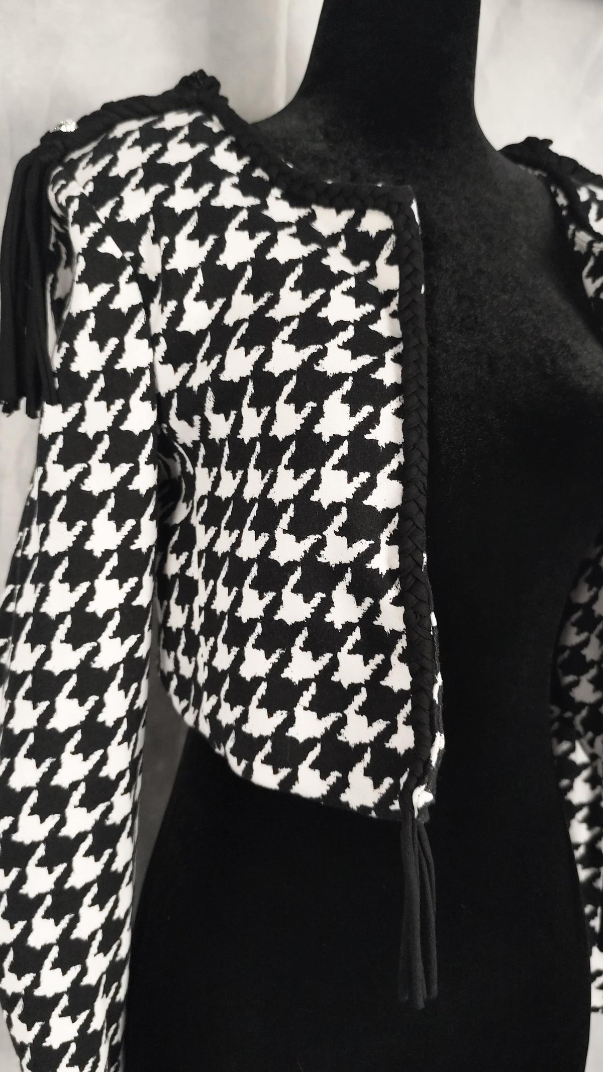A black and white houndstooth cropped jacket with black fringe and a black braided trim, displayed on a black mannequin torso.