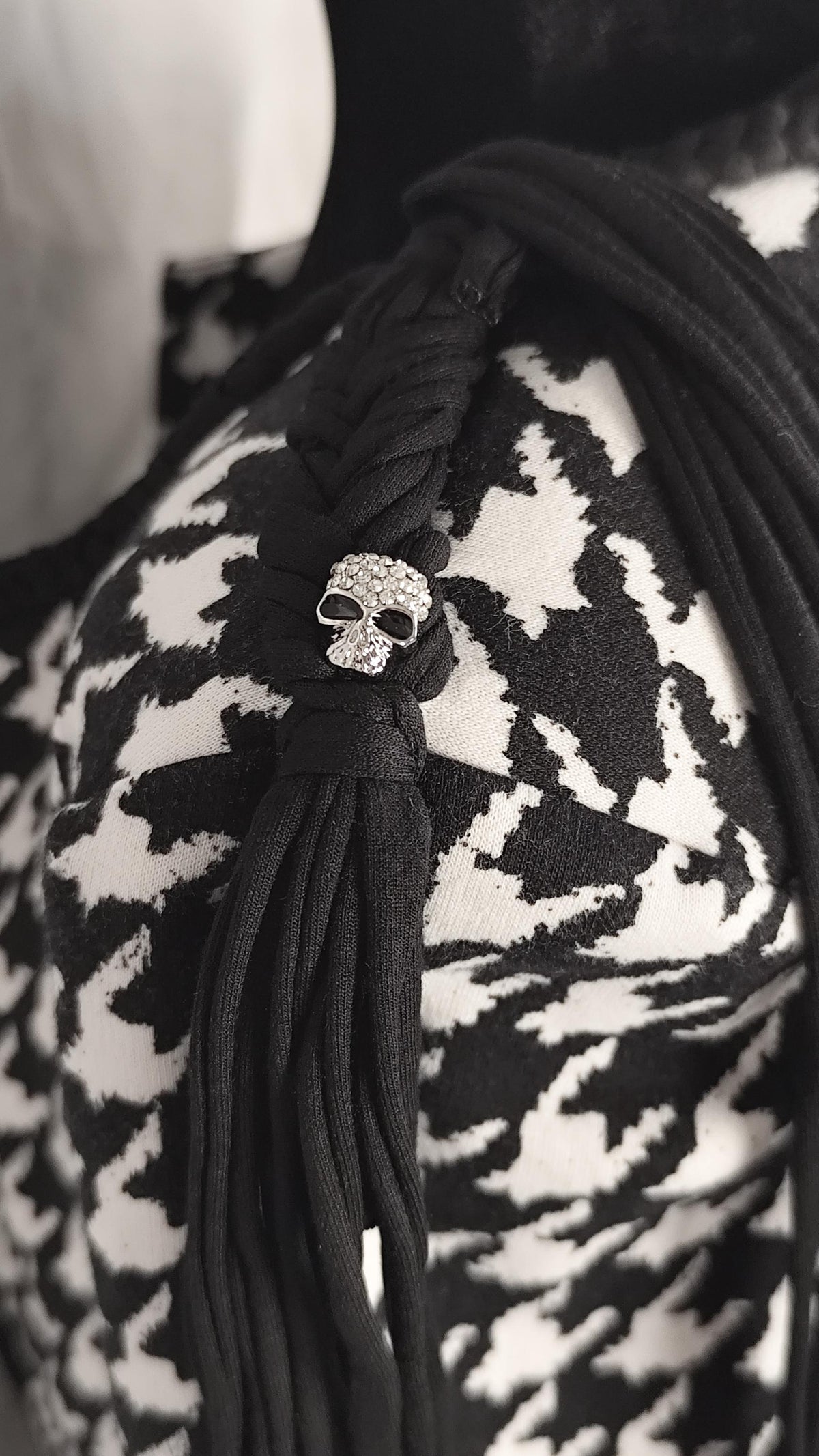 A close-up of a black textile waste braid accessory with fringe and a small skull charm covered in clear rhinestones, worn over a black-and-white patterned top.